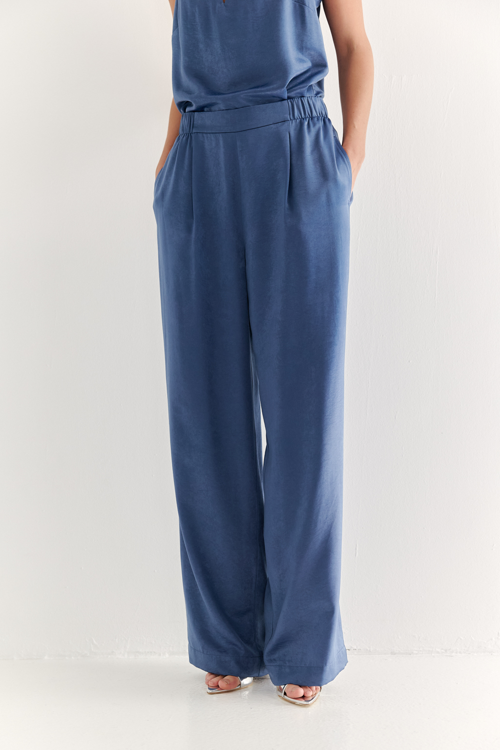 Брюки, широкие и прямые по всей длине, выполнены из струящегося атласа, что является воплощением актуального тренда на мягкий комфорт. Стильный цвет классического денима и фактура ткани, напоминающая мокрый шёлк, определяют ключевое направление в моде: создание атмосферы комфорта, пронизанной заботой и бережным отношением к себе. Брюки оснащены удобными карманами и эластичным поясом, обеспечивающими максимальный комфорт и удобство. Идеальным дополнением к этим брюкам станут топ и жакет в пижамном стиле, позволяющие создать элегантный монохромный образ.