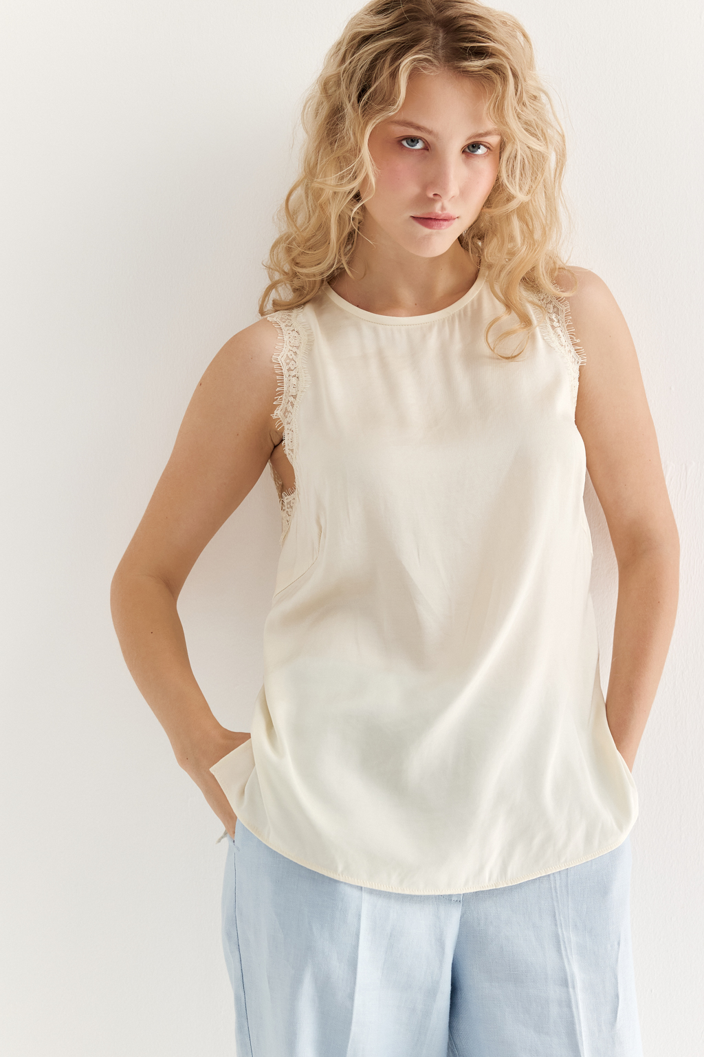 Блуза без рукавов из струящегося сатина в оттенке сливочное масло. Круглая горловина обработана кантом, на спинке воздушная петля с застежкой на пуговицу, по проймам настрочена тесьма из гипюра в тон основной ткани.
