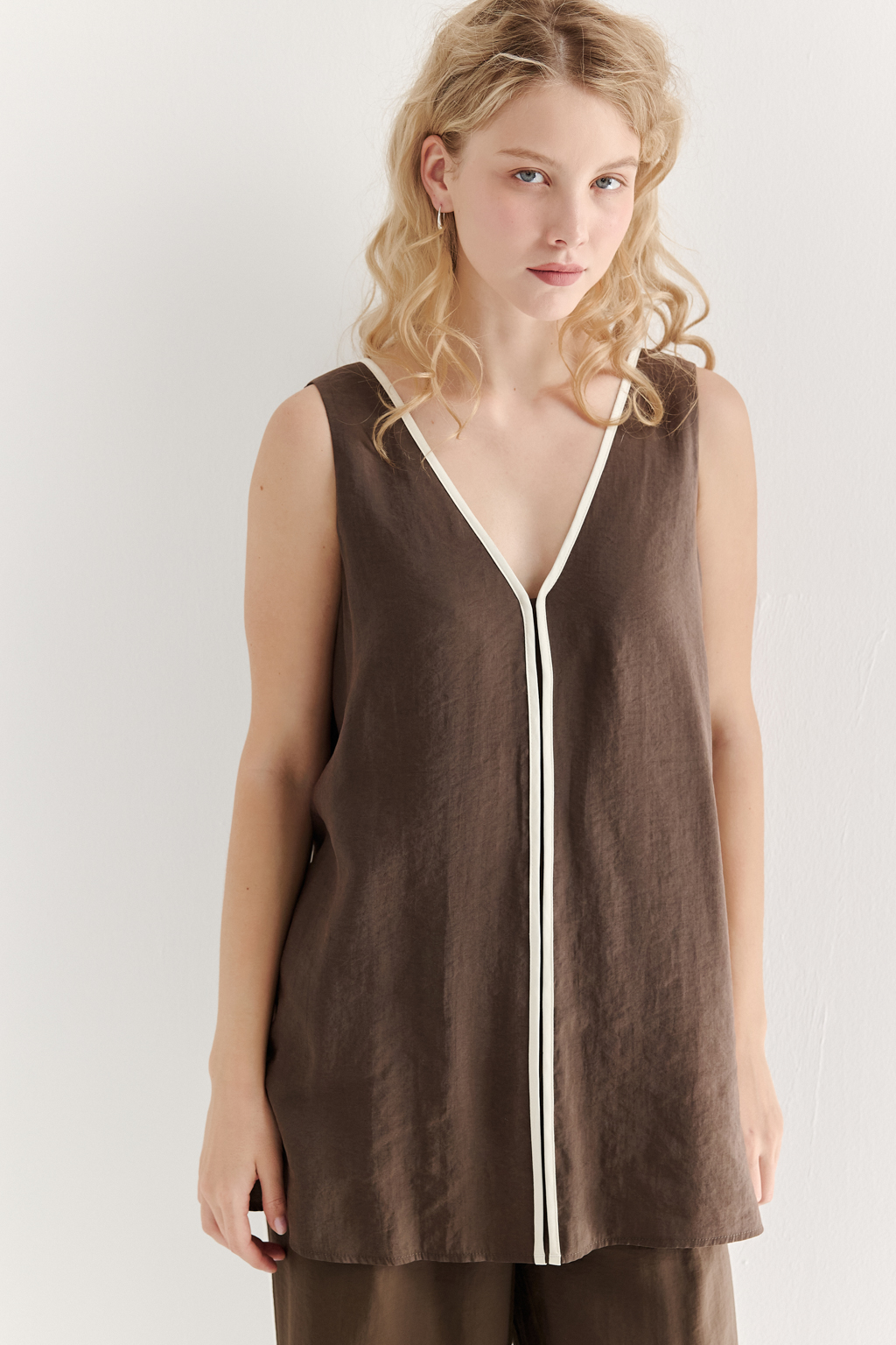 Блуза без рукавов, трапециевидного силуэта, выполнена из модала насыщенного шоколадно-коричневого оттенка и отделана контрастным кантом. Ткань обладает бархатистой текстурой, создающей так называемый "Peach эффект": благодаря специальной обработке на поверхности ткани образуется деликатный ворс, тактильно напоминающий кожицу персика. Горловина, центральный шов переда, боковые швы, а также шнуровка на спинке оформлены шелковистым кантом нежного молочного цвета. В боковых швах предусмотрены разрезы. Данная блуза прекрасно сочетается с элегантными брюками с лампасами из аналогичной ткани