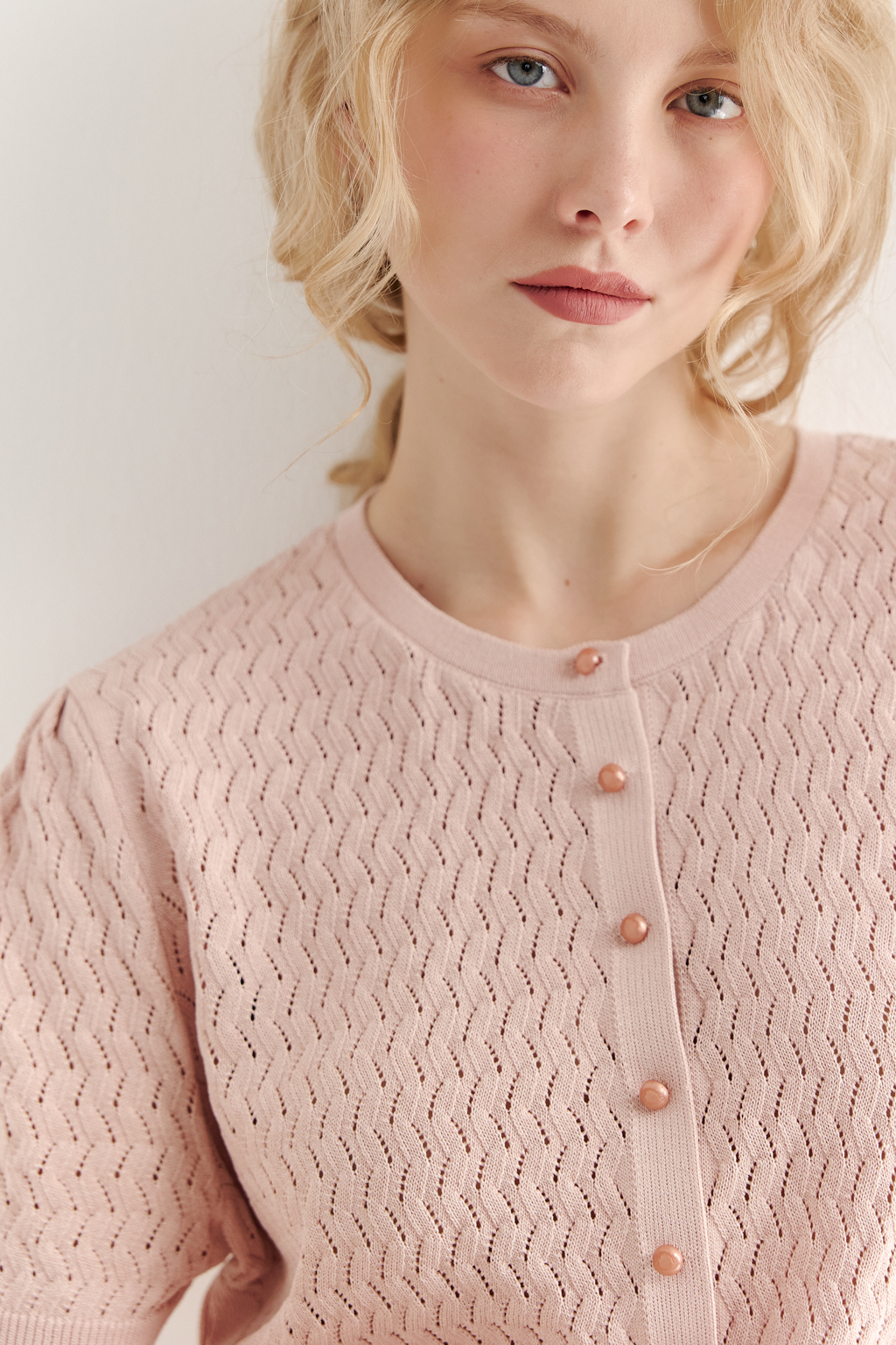 Джемпер нежно-розового цвета, связанный из тонкой тактильной пряжи, в составе которой вискоза и нейлон. В дизайне джемпера применен принцип контраста фактур, в данном случае – полупрозрачного ажура и ластика. Круглая горловина и планка с застежкой на 9 пуговиц-жемчужин, рукав длиной одна вторая.