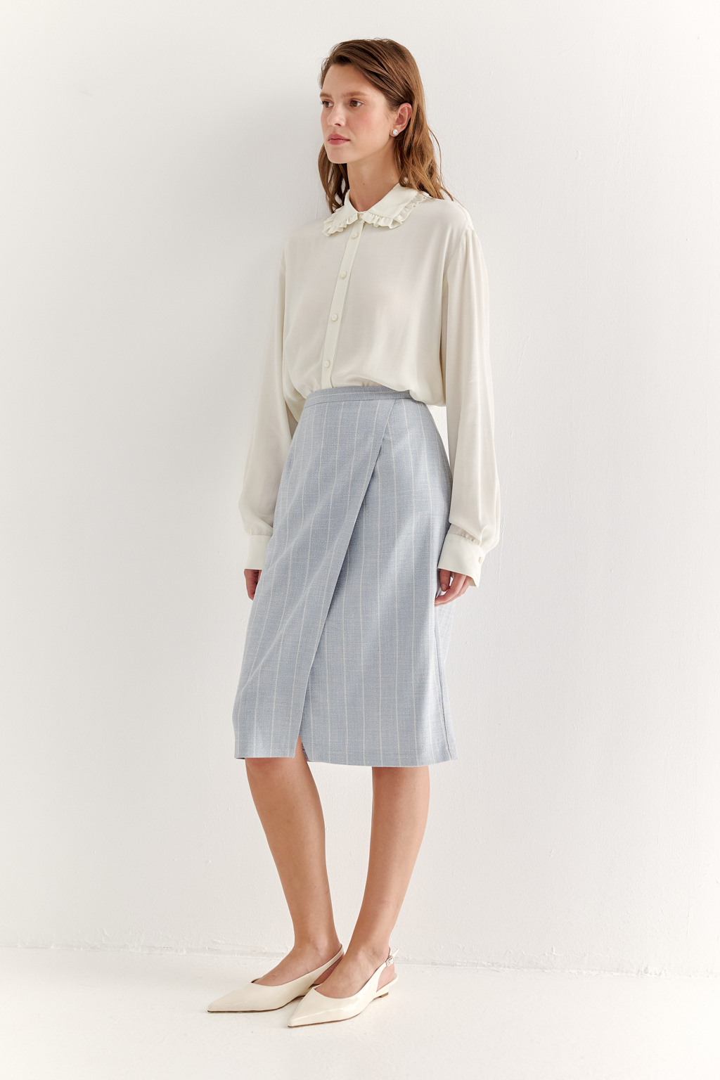 Прямая юбка с запахом выполнена из ткани, представляющей собой уникальную разработку бренда. Сочетание волокон хлопка, вискозы, полиэстера и эластана обеспечивает максимальный комфорт и практичность изделия. Нежный голубой цвет  эффектно подчёркнут светлой полоской в широком раппорте. Юбка идеально сочетается с жакетом архитектурного кроя из той же ткани, создавая завершённый формальный образ.