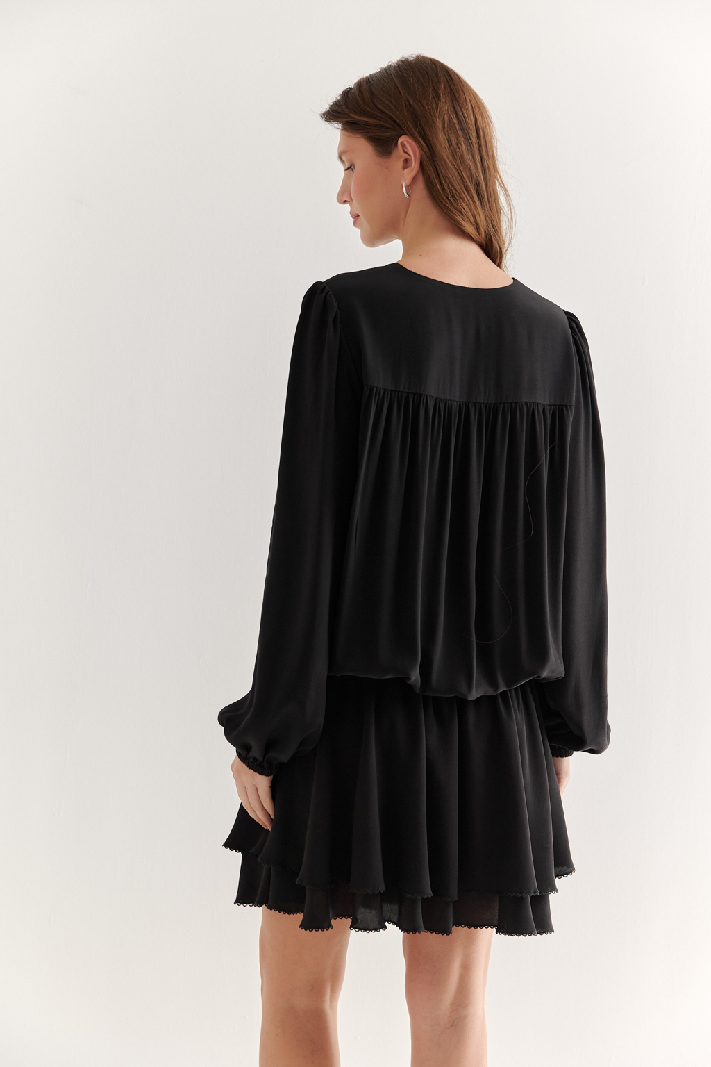 Платье, из струящейся креповой вискозы в черном цвете, представляет собой воплощение модных тенденций, умело играя с объемами с акцентом на заниженную линию талии. Это короткое, воздушное и исполненное романтики платье, сознательно избегающее вычурных деталей, является идеальным решением для создания актуального летнего городского образа. Объем платья образуется благодаря изящным сборкам, расположенным по кокеткам переда и спинки. Отрезная юбка, расширяющаяся к низу, украшена деликатной декоративной строчкой-вышивкой в виде фестонов. Линия отреза юбки оформлена эластичной тесьмой-стрейч, что дает возможность регулировать длину. Платье гармонично сочетается с рубашками или жакетами, предоставляя свободу для экспериментов со стилем. При желании можно усложнить образ образа с помощью леггинсов и также подчеркнуть талию элегантным кожаным ремнем.