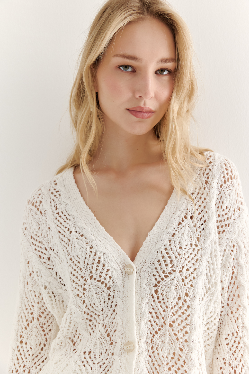 Белый вязаный кардиган оверсайз силуэта из хлопковой пряжи. Кардиган связан в сложной технике комбинирования сетки и ажурных ромбов. Рукав прямой удлиненный, V-образный вырез, борт отделан планкой, застежка по борту на 3 пуговицы, низ кардигана и рукав – сложные фестоны.