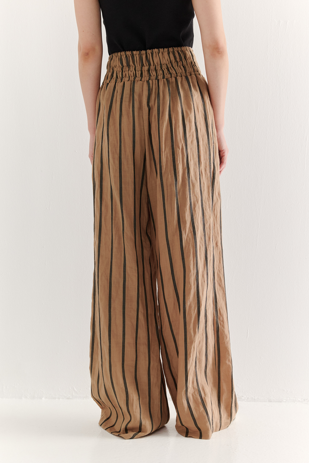 Брюки прямого кроя с широким поясом, собранным в сборку, сшиты из платьевой ткани в тонкую полоску. Состав ткани - сочетание волокон крапивы и хлопка, обработанных с эффектом растительного окрашивания (natural dye). Темная полоска на бежевом фоне идеально гармонирует с силуэтом широких брюк, подчеркивая их элегантную простоту. Пояс брюк широкий, собран на три ряда кулисок с резинками, что позволяет регулировать его высоту посадки относительно линии талии. Ткань обладает свойством создавать естественный крэш-эффект, придающий изделию легкую текстурную небрежность и объем, при этом брюки не требуют сложного ухода.