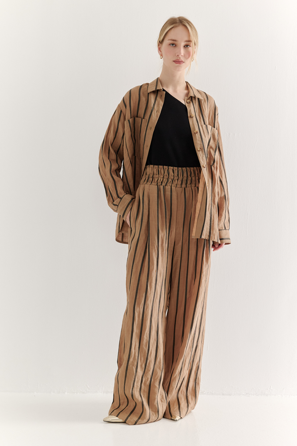 Блуза-рубашка свободного кроя изготовлена сшиты из ткани в темную полоску на песочном фоне. Ткань сочетает в себе волокна крапивы и хлопка, прошедшие обработку с использованием натуральных красителей (natural dye). Дизайн, вдохновлённый классической мужской рубашкой, включает в себя воротник-стойку, планку с застёжкой и длинные рукава с манжетами. Свободный расслабленный стиль рубашки идеально рифмуется с комфортными брюками из этой же ткани и компактными трикотажными джемперами и топами. Яркое решение образа создает комфортный и запоминающийся образ