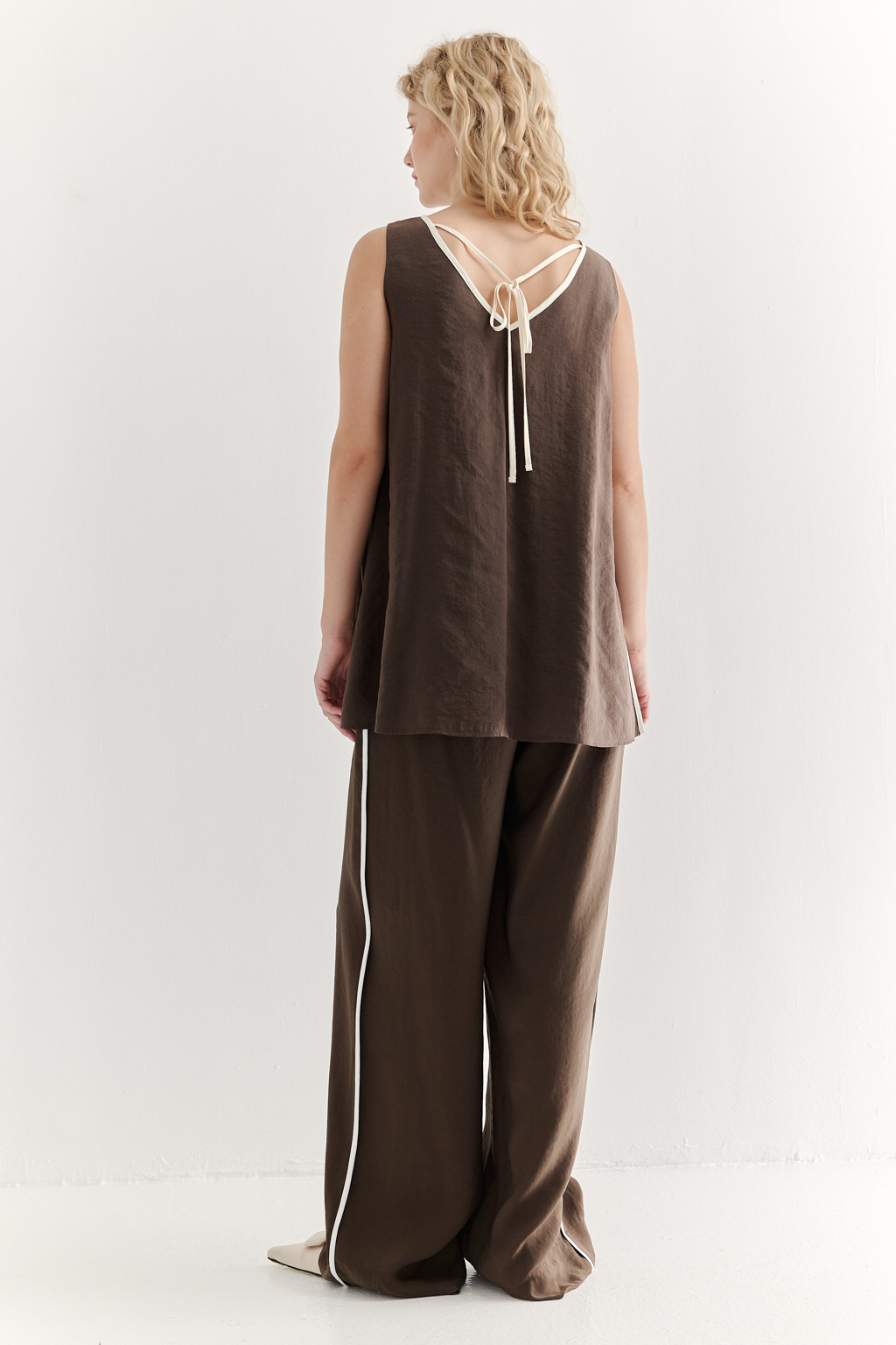 Блуза без рукавов, трапециевидного силуэта, выполнена из модала насыщенного шоколадно-коричневого оттенка и отделана контрастным кантом. Ткань обладает бархатистой текстурой, создающей так называемый "Peach эффект": благодаря специальной обработке на поверхности ткани образуется деликатный ворс, тактильно напоминающий кожицу персика. Горловина, центральный шов переда, боковые швы, а также шнуровка на спинке оформлены шелковистым кантом нежного молочного цвета. В боковых швах предусмотрены разрезы. Данная блуза прекрасно сочетается с элегантными брюками с лампасами из аналогичной ткани