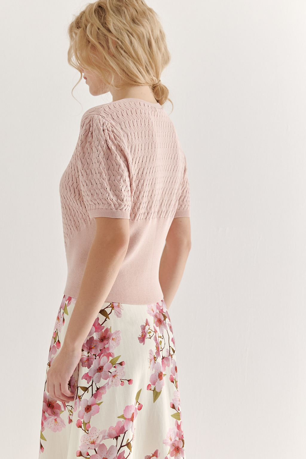 Джемпер нежно-розового цвета, связанный из тонкой тактильной пряжи, в составе которой вискоза и нейлон. В дизайне джемпера применен принцип контраста фактур, в данном случае – полупрозрачного ажура и ластика. Круглая горловина и планка с застежкой на 9 пуговиц-жемчужин, рукав длиной одна вторая.