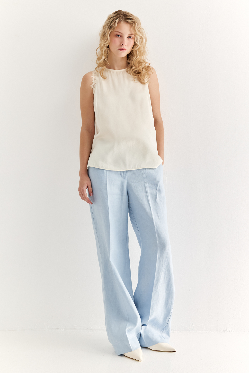Блуза без рукавов из струящегося сатина в оттенке сливочное масло. Круглая горловина обработана кантом, на спинке воздушная петля с застежкой на пуговицу, по проймам настрочена тесьма из гипюра в тон основной ткани.