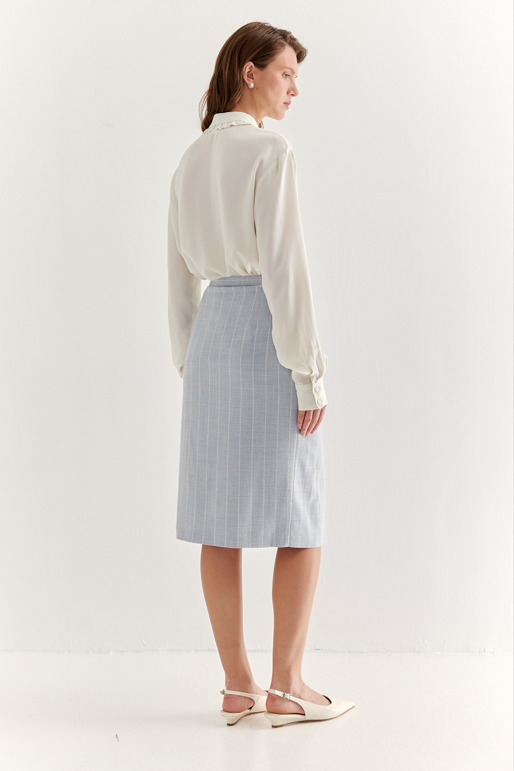 Прямая юбка с запахом выполнена из ткани, представляющей собой уникальную разработку бренда. Сочетание волокон хлопка, вискозы, полиэстера и эластана обеспечивает максимальный комфорт и практичность изделия. Нежный голубой цвет  эффектно подчёркнут светлой полоской в широком раппорте. Юбка идеально сочетается с жакетом архитектурного кроя из той же ткани, создавая завершённый формальный образ.