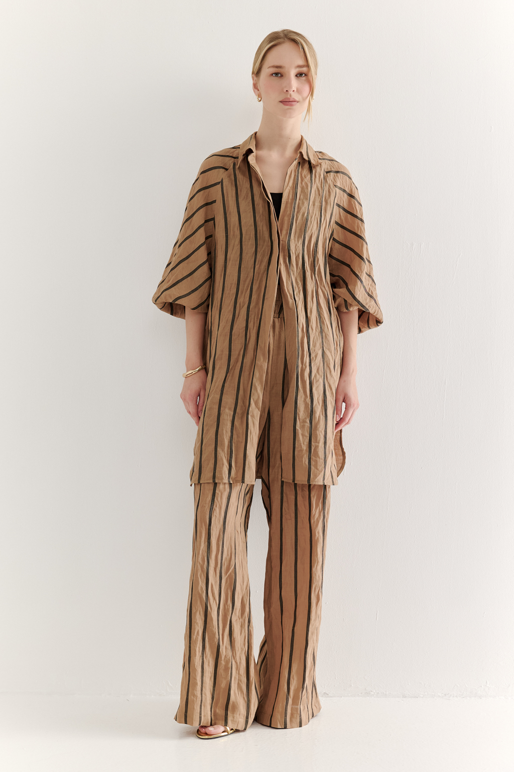 Блуза-туника сшита из платьевой ткани в тонкую полоску, состав которой представляет собой натуральное сочетание волокон крапивы и хлопка с эффектом растительного окрашивания (natural dye). Модель прямого кроя оснащена супатной застежкой на пуговицах, рубашечным воротником-стойкой и объемными рукавами-реглан длиной одна вторая. Ткань позволяет создать естественный крэш-эффект, придающий изделию легкую текстурную небрежность, при этом блуза не требует сложного ухода. Для сохранения формы рекомендуется хранить блузу в свернутом состоянии.