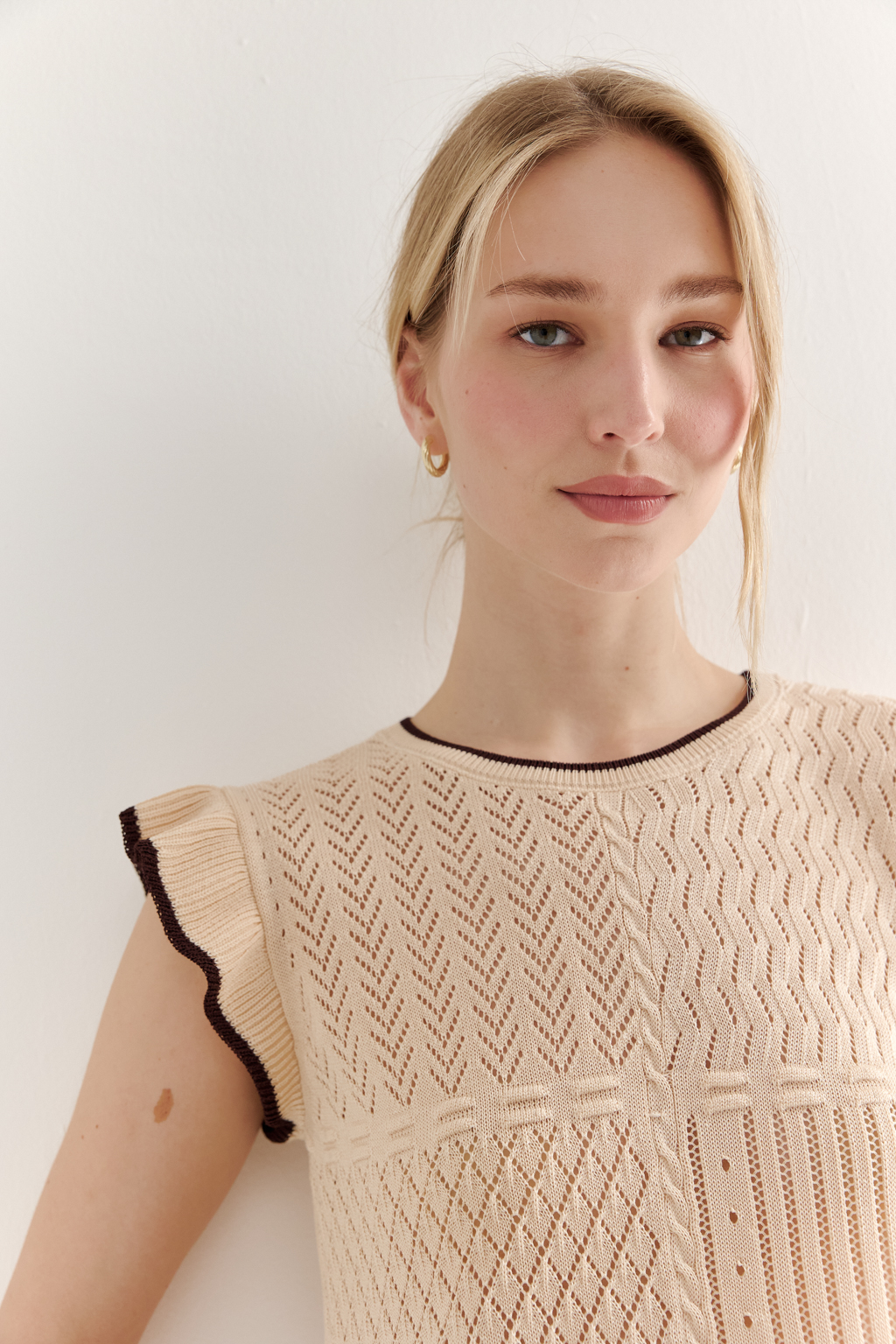 Кремовый топ в стиле пэчворк, в котором соединились четыре техники ажурной вязки, круглая горловина и деликатные рюши по пройме. Контрастная отделка коричневого цвета расположена по линиям горловины, низа и краю рюши.