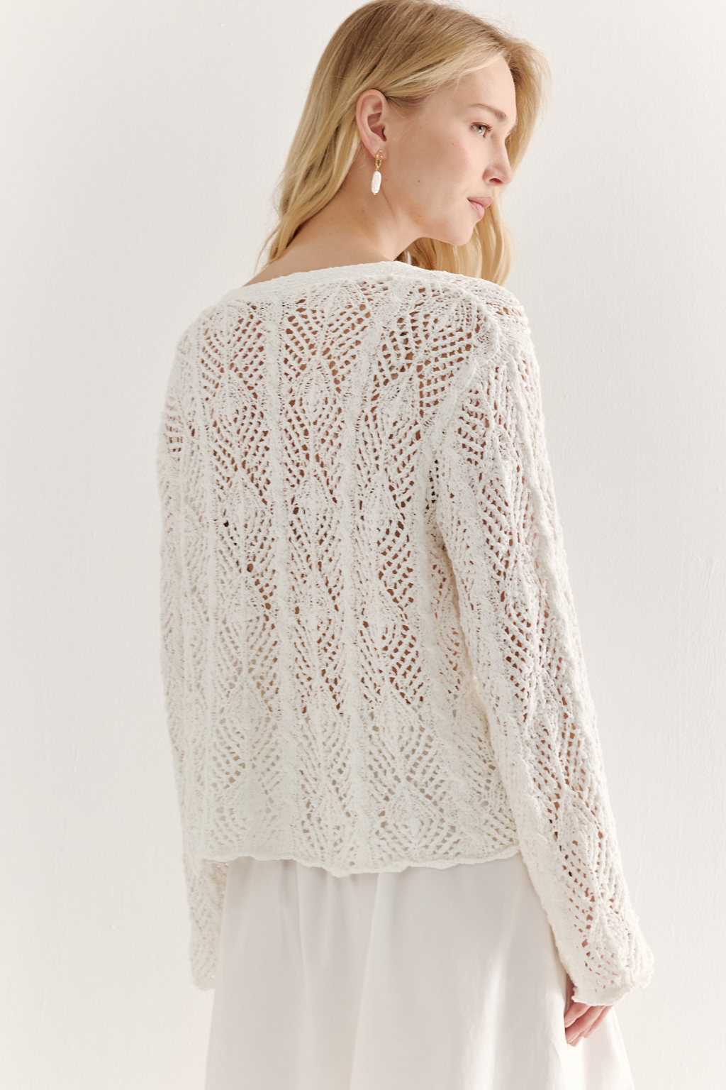Белый вязаный кардиган оверсайз силуэта из хлопковой пряжи. Кардиган связан в сложной технике комбинирования сетки и ажурных ромбов. Рукав прямой удлиненный, V-образный вырез, борт отделан планкой, застежка по борту на 3 пуговицы, низ кардигана и рукав – сложные фестоны.