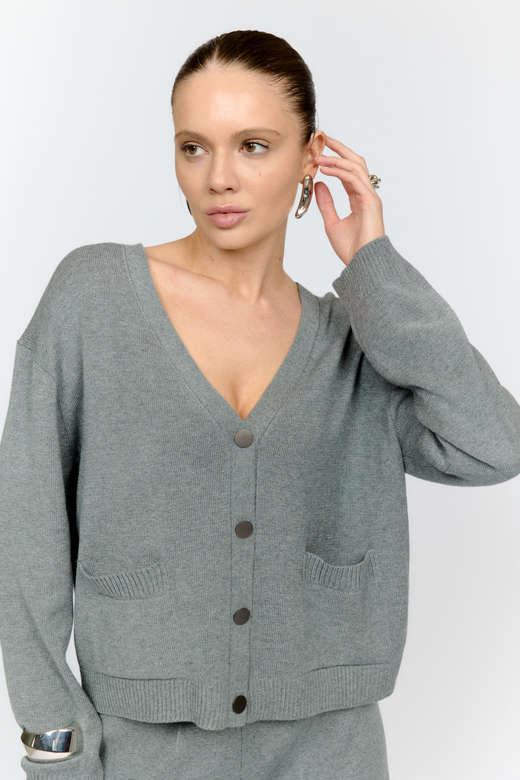 Трикотажный кардиган в сером цвете с V-образным вырезом горловины, на пуговицах, со втачным рукавом, двумя накладными карманами на полочке. Можно использовать в качестве альтернативы жакетам и носить вторым слоем в расстегнутом виде на более тонкие верха.