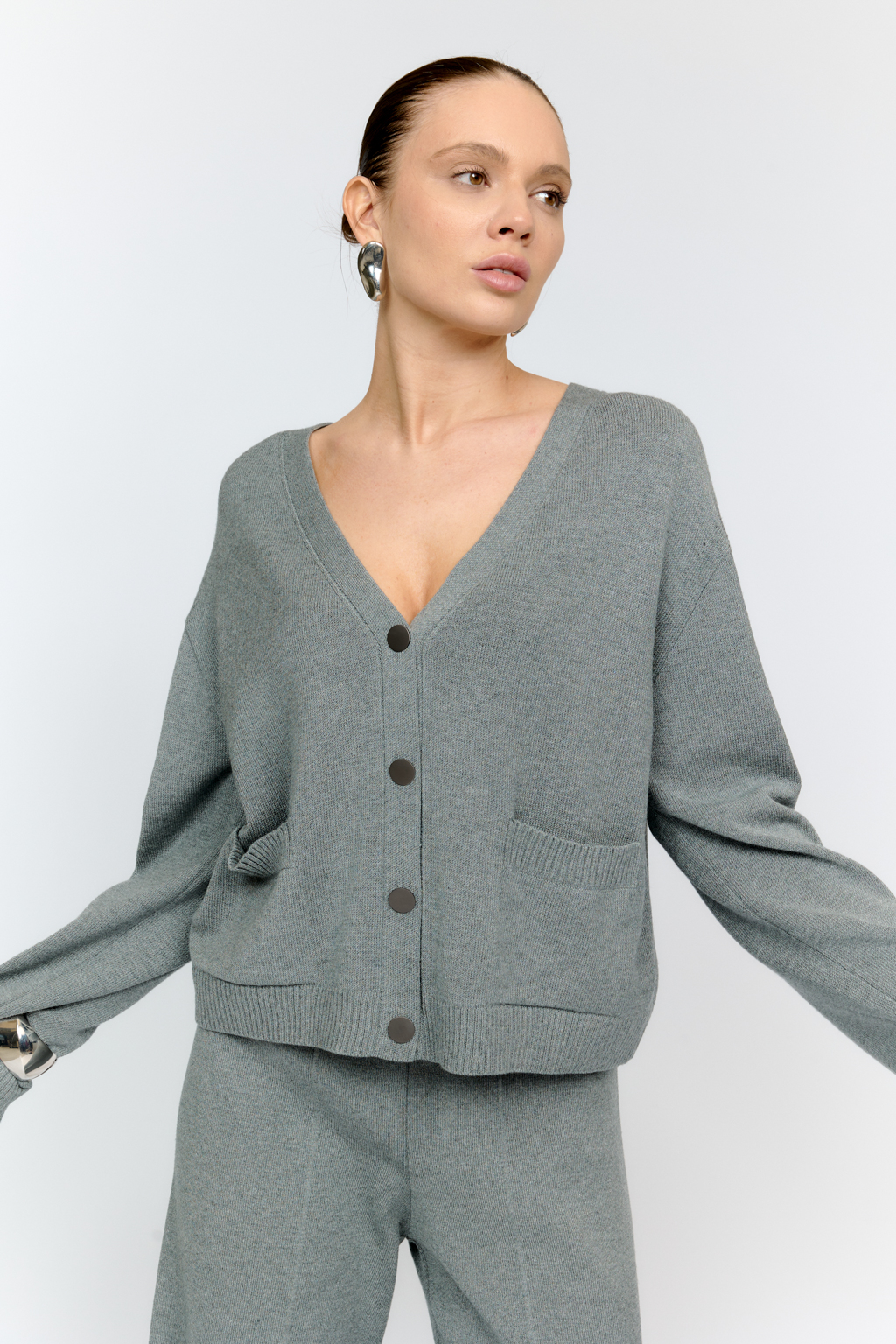 Трикотажный кардиган в сером цвете с V-образным вырезом горловины, на пуговицах, со втачным рукавом, двумя накладными карманами на полочке. Можно использовать в качестве альтернативы жакетам и носить вторым слоем в расстегнутом виде на более тонкие верха.