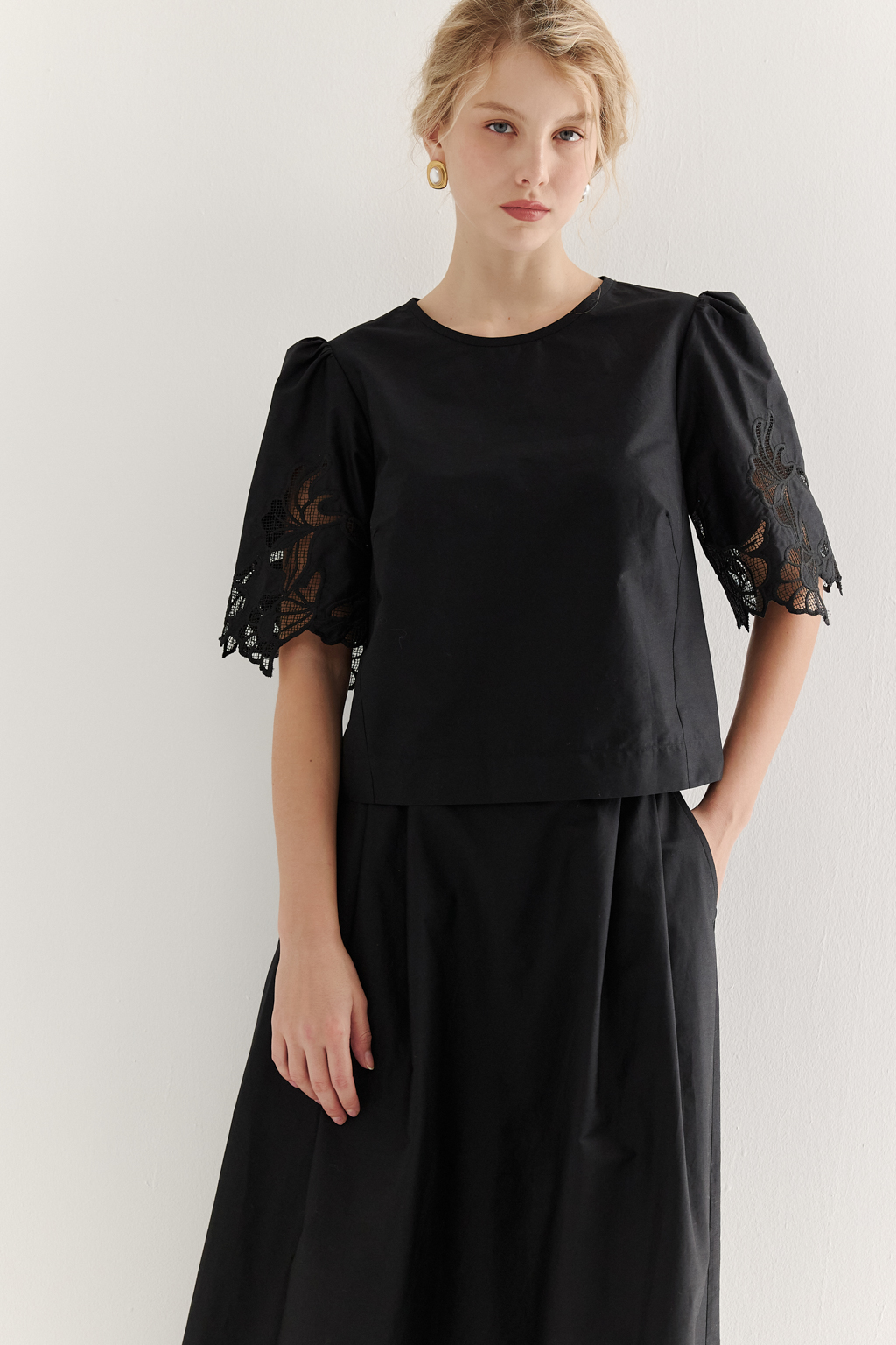 Блуза-кроп выполненная из хлопкового полотна, декорирована нежным шитьем-решелье черного цвета. Укороченный силуэт блузы дополнен широкой подгибкой по низу и элегантными разрезами в боковых швах. Горловина оформлена в виде лодочки, визуально удлиняя шею. Широкие рукава имеют сборку по окату и украшены шитьем по низу, что придает образу легкости и женственности. Молния, расположенная в среднем шве спинки, обеспечивает комфорт и удобство при носке. Идеально подойдет к юбке с шитьем-решелье белого цвета.