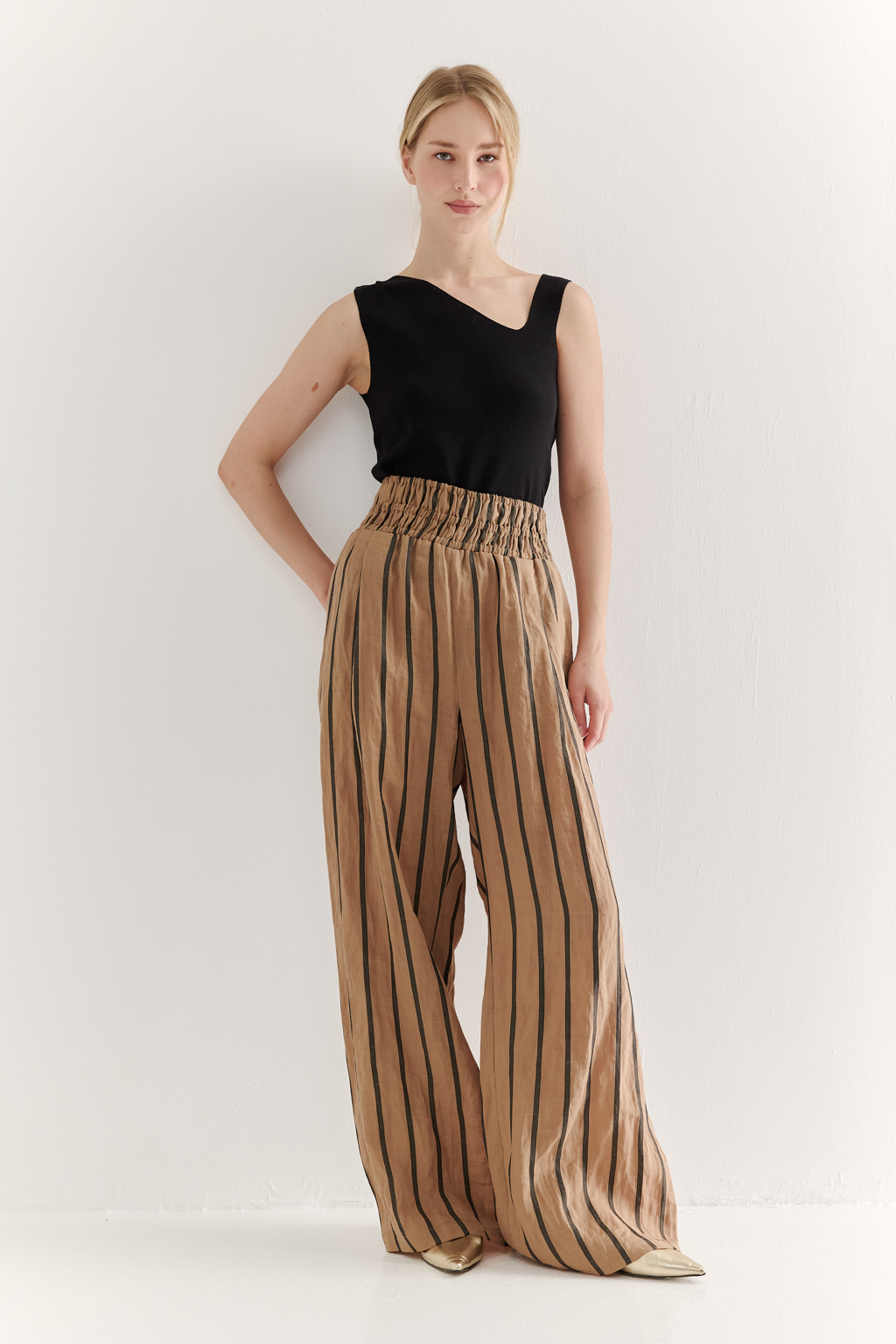 Брюки прямого кроя с широким поясом, собранным в сборку, сшиты из платьевой ткани в тонкую полоску. Состав ткани - сочетание волокон крапивы и хлопка, обработанных с эффектом растительного окрашивания (natural dye). Темная полоска на бежевом фоне идеально гармонирует с силуэтом широких брюк, подчеркивая их элегантную простоту. Пояс брюк широкий, собран на три ряда кулисок с резинками, что позволяет регулировать его высоту посадки относительно линии талии. Ткань обладает свойством создавать естественный крэш-эффект, придающий изделию легкую текстурную небрежность и объем, при этом брюки не требуют сложного ухода.
