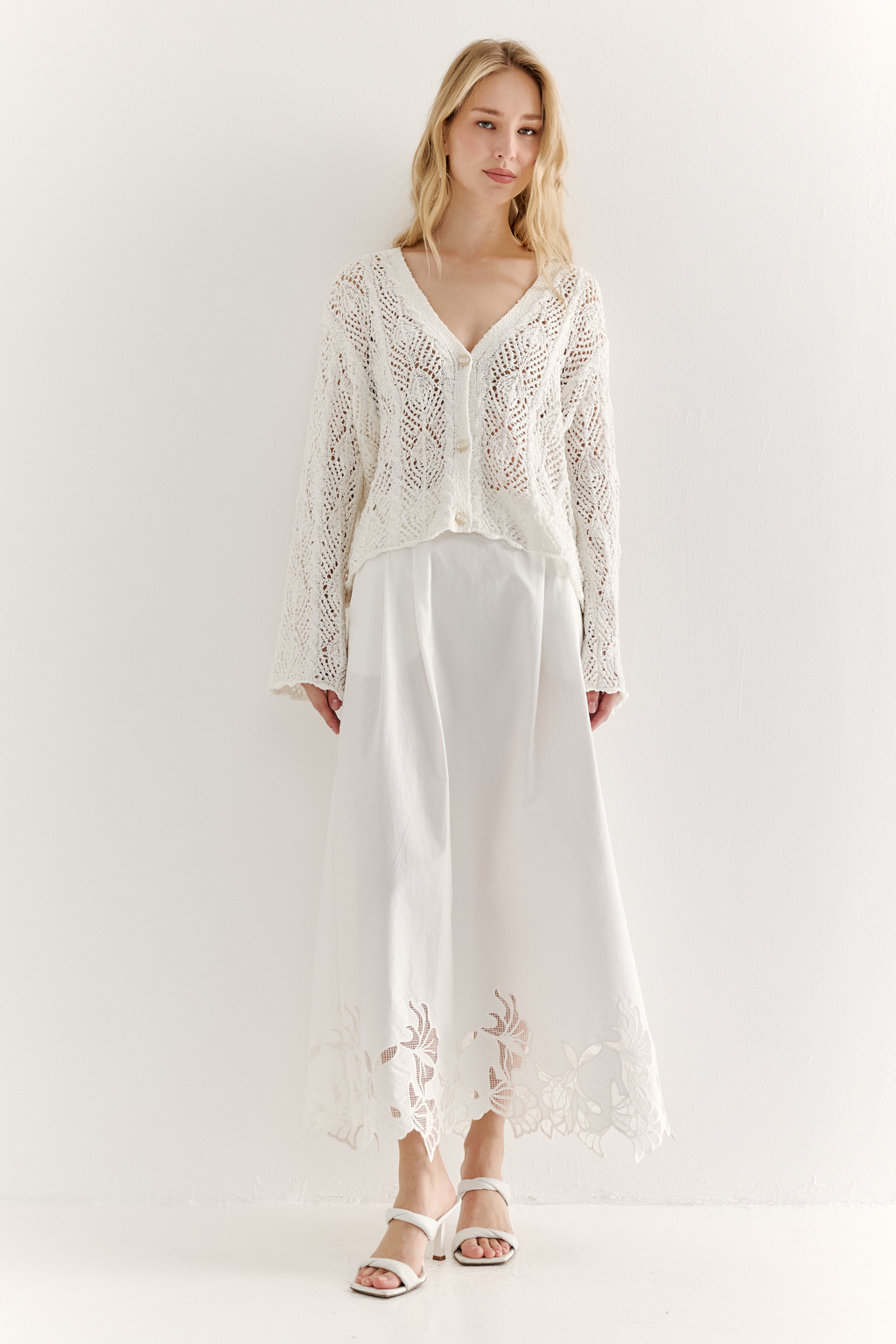 Белый вязаный кардиган оверсайз силуэта из хлопковой пряжи. Кардиган связан в сложной технике комбинирования сетки и ажурных ромбов. Рукав прямой удлиненный, V-образный вырез, борт отделан планкой, застежка по борту на 3 пуговицы, низ кардигана и рукав – сложные фестоны.