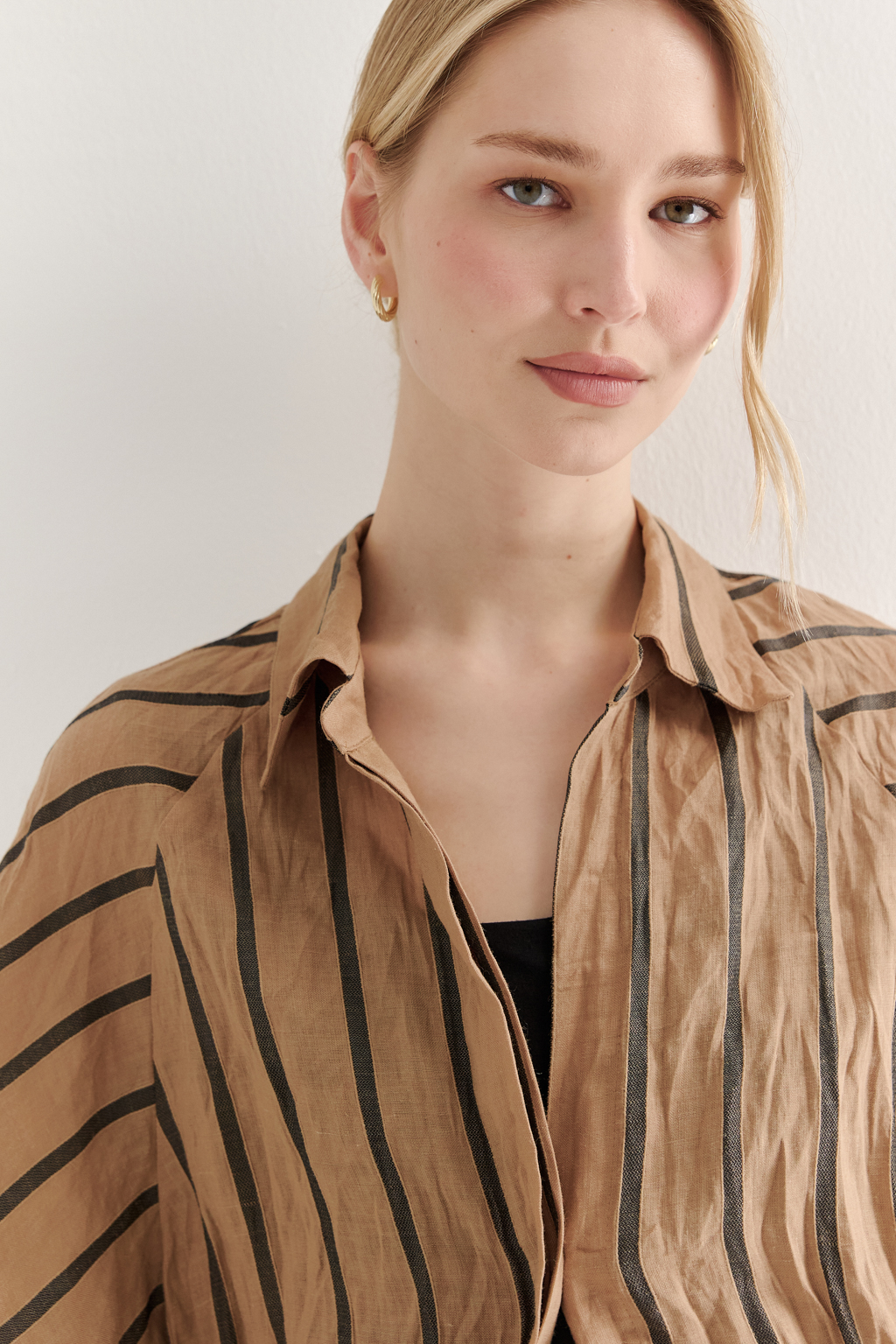 Блуза-туника сшита из платьевой ткани в тонкую полоску, состав которой представляет собой натуральное сочетание волокон крапивы и хлопка с эффектом растительного окрашивания (natural dye). Модель прямого кроя оснащена супатной застежкой на пуговицах, рубашечным воротником-стойкой и объемными рукавами-реглан длиной одна вторая. Ткань позволяет создать естественный крэш-эффект, придающий изделию легкую текстурную небрежность, при этом блуза не требует сложного ухода. Для сохранения формы рекомендуется хранить блузу в свернутом состоянии.