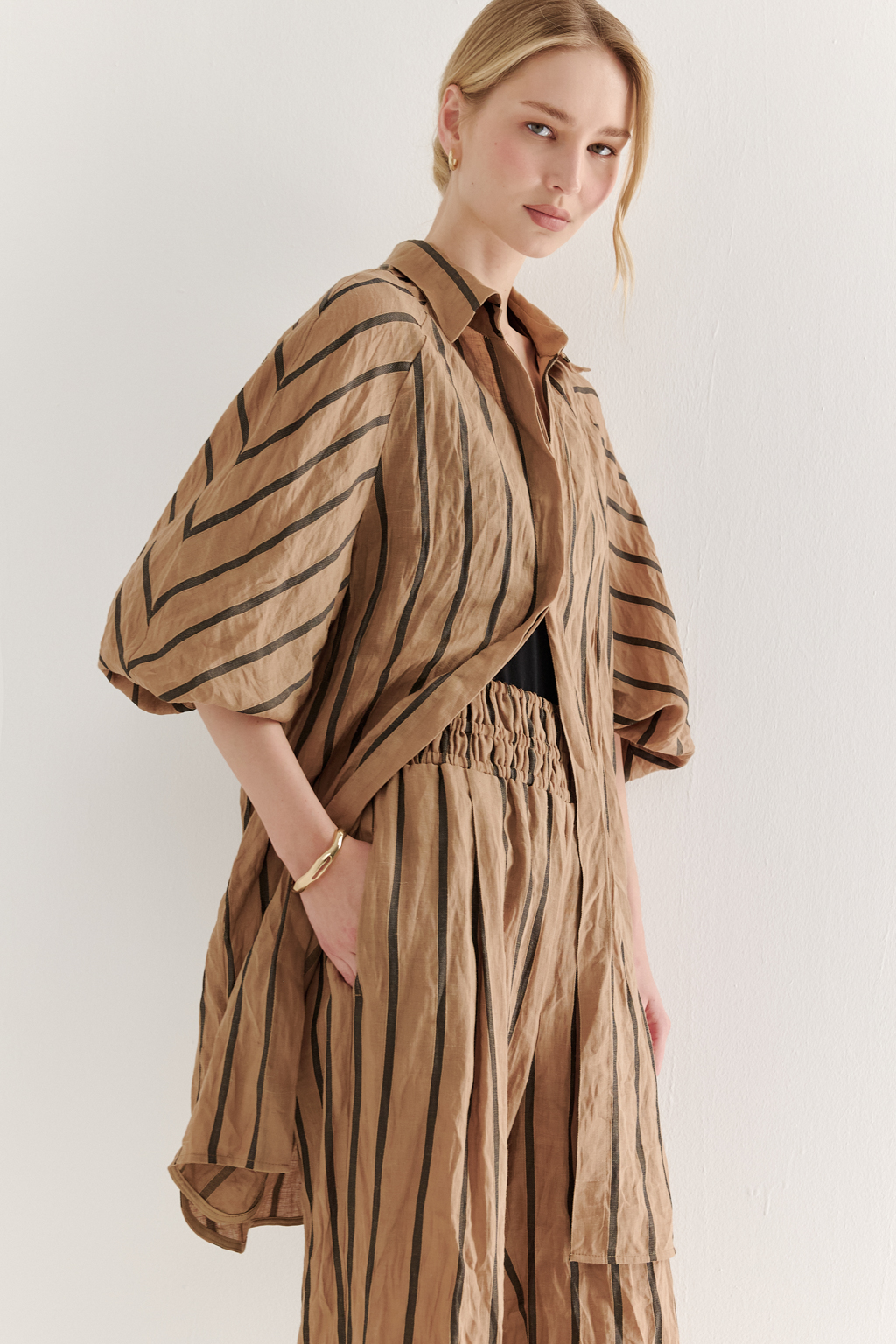 Брюки прямого кроя с широким поясом, собранным в сборку, сшиты из платьевой ткани в тонкую полоску. Состав ткани - сочетание волокон крапивы и хлопка, обработанных с эффектом растительного окрашивания (natural dye). Темная полоска на бежевом фоне идеально гармонирует с силуэтом широких брюк, подчеркивая их элегантную простоту. Пояс брюк широкий, собран на три ряда кулисок с резинками, что позволяет регулировать его высоту посадки относительно линии талии. Ткань обладает свойством создавать естественный крэш-эффект, придающий изделию легкую текстурную небрежность и объем, при этом брюки не требуют сложного ухода.