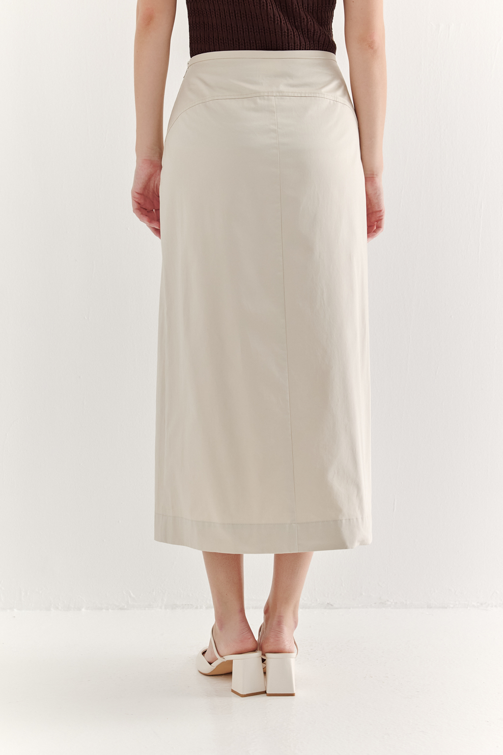 Изысканная юбка-миди А-силуэта выполнена из полированного хлопка нежного светло-бежевого оттенка. Передняя часть юбки украшена тремя  складками, искусно формирующими силуэт. В крайней левой складке расположена шлица, прикрывающая высокий разрез.