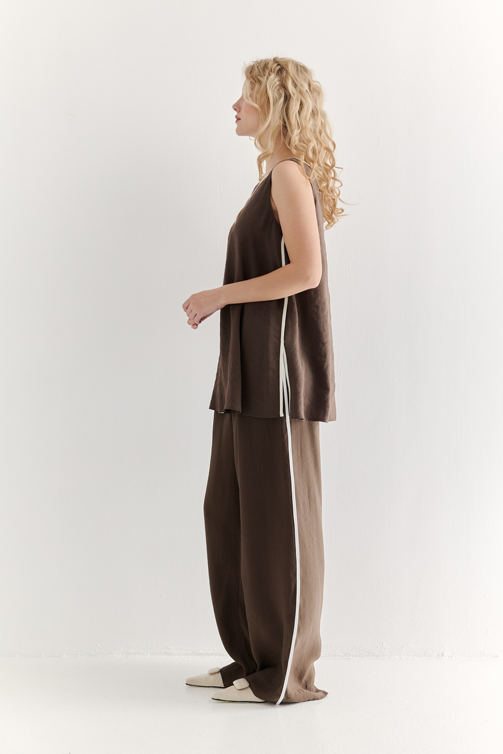 Блуза без рукавов, трапециевидного силуэта, выполнена из модала насыщенного шоколадно-коричневого оттенка и отделана контрастным кантом. Ткань обладает бархатистой текстурой, создающей так называемый "Peach эффект": благодаря специальной обработке на поверхности ткани образуется деликатный ворс, тактильно напоминающий кожицу персика. Горловина, центральный шов переда, боковые швы, а также шнуровка на спинке оформлены шелковистым кантом нежного молочного цвета. В боковых швах предусмотрены разрезы. Данная блуза прекрасно сочетается с элегантными брюками с лампасами из аналогичной ткани