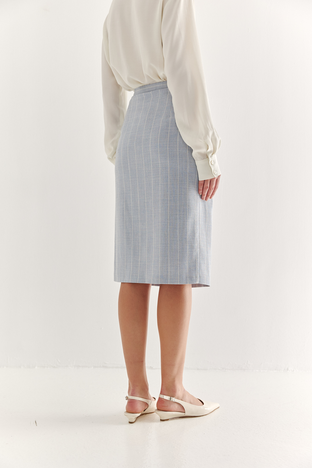 Прямая юбка с запахом выполнена из ткани, представляющей собой уникальную разработку бренда. Сочетание волокон хлопка, вискозы, полиэстера и эластана обеспечивает максимальный комфорт и практичность изделия. Нежный голубой цвет  эффектно подчёркнут светлой полоской в широком раппорте. Юбка идеально сочетается с жакетом архитектурного кроя из той же ткани, создавая завершённый формальный образ.