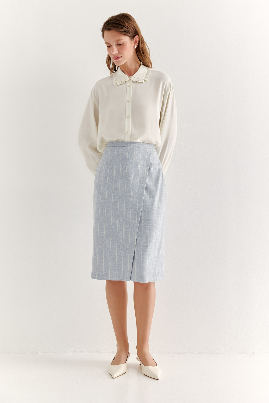 Прямая юбка с запахом выполнена из ткани, представляющей собой уникальную разработку бренда. Сочетание волокон хлопка, вискозы, полиэстера и эластана обеспечивает максимальный комфорт и практичность изделия. Нежный голубой цвет  эффектно подчёркнут светлой полоской в широком раппорте. Юбка идеально сочетается с жакетом архитектурного кроя из той же ткани, создавая завершённый формальный образ.