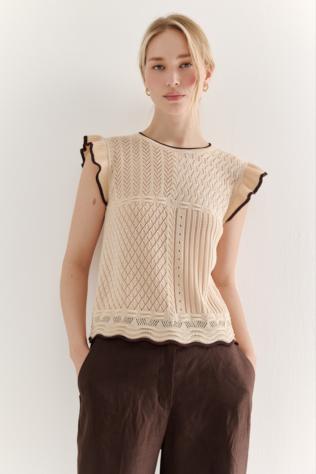 Кремовый топ в стиле пэчворк, в котором соединились четыре техники ажурной вязки, круглая горловина и деликатные рюши по пройме. Контрастная отделка коричневого цвета расположена по линиям горловины, низа и краю рюши.