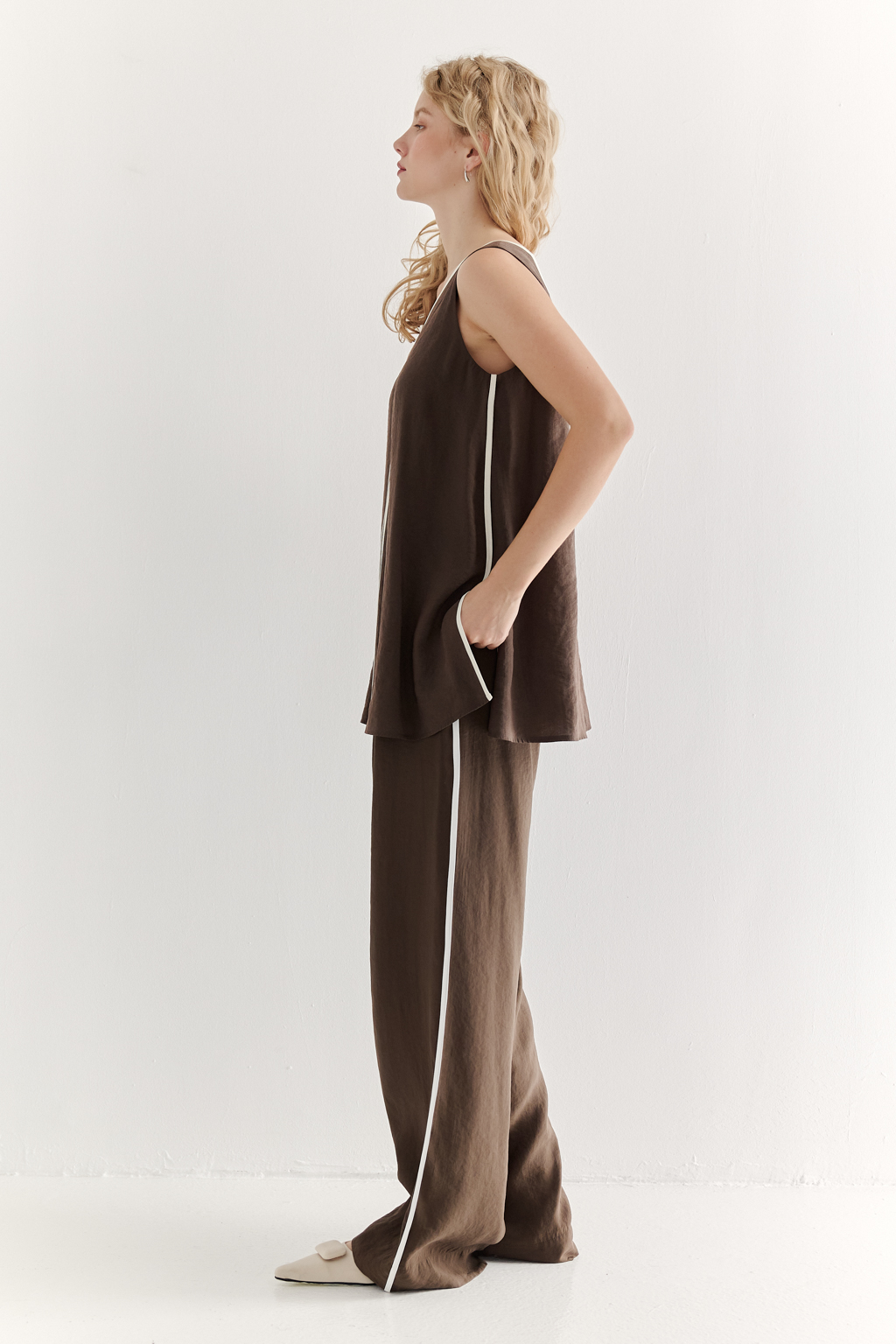 Блуза без рукавов, трапециевидного силуэта, выполнена из модала насыщенного шоколадно-коричневого оттенка и отделана контрастным кантом. Ткань обладает бархатистой текстурой, создающей так называемый "Peach эффект": благодаря специальной обработке на поверхности ткани образуется деликатный ворс, тактильно напоминающий кожицу персика. Горловина, центральный шов переда, боковые швы, а также шнуровка на спинке оформлены шелковистым кантом нежного молочного цвета. В боковых швах предусмотрены разрезы. Данная блуза прекрасно сочетается с элегантными брюками с лампасами из аналогичной ткани