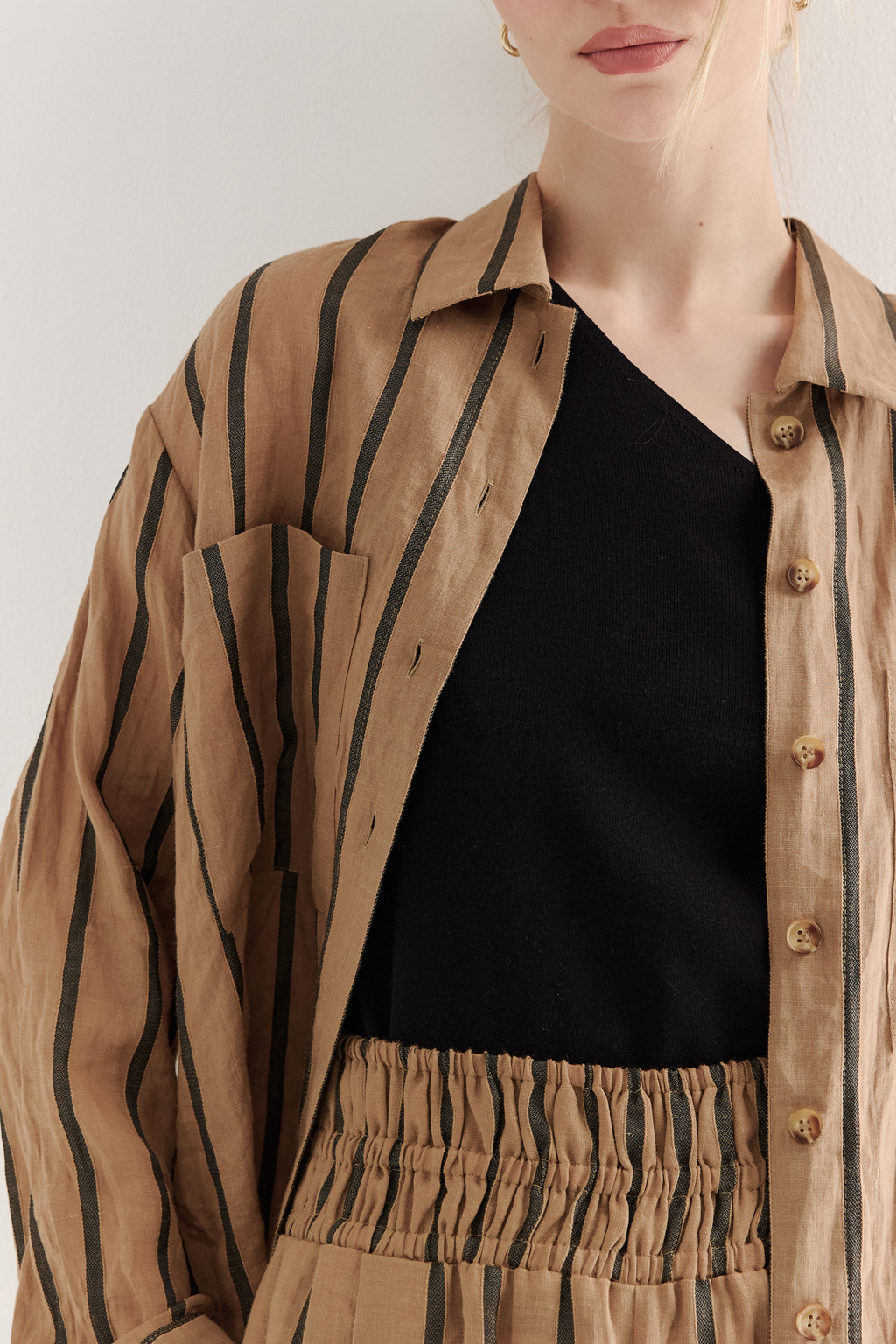 Блуза-рубашка свободного кроя изготовлена сшиты из ткани в темную полоску на песочном фоне. Ткань сочетает в себе волокна крапивы и хлопка, прошедшие обработку с использованием натуральных красителей (natural dye). Дизайн, вдохновлённый классической мужской рубашкой, включает в себя воротник-стойку, планку с застёжкой и длинные рукава с манжетами. Свободный расслабленный стиль рубашки идеально рифмуется с комфортными брюками из этой же ткани и компактными трикотажными джемперами и топами. Яркое решение образа создает комфортный и запоминающийся образ