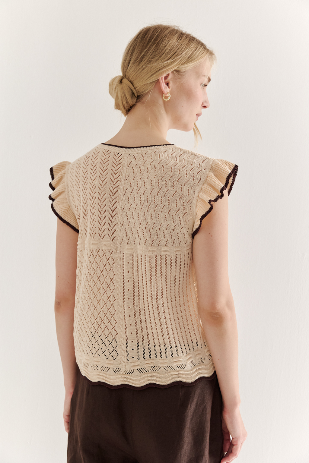 Кремовый топ в стиле пэчворк, в котором соединились четыре техники ажурной вязки, круглая горловина и деликатные рюши по пройме. Контрастная отделка коричневого цвета расположена по линиям горловины, низа и краю рюши.
