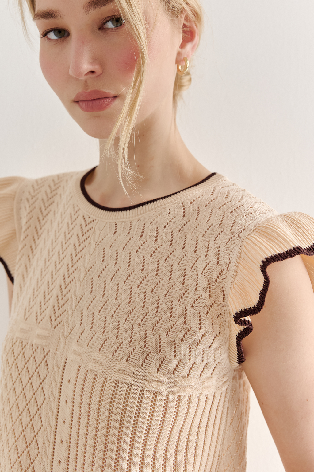 Кремовый топ в стиле пэчворк, в котором соединились четыре техники ажурной вязки, круглая горловина и деликатные рюши по пройме. Контрастная отделка коричневого цвета расположена по линиям горловины, низа и краю рюши.
