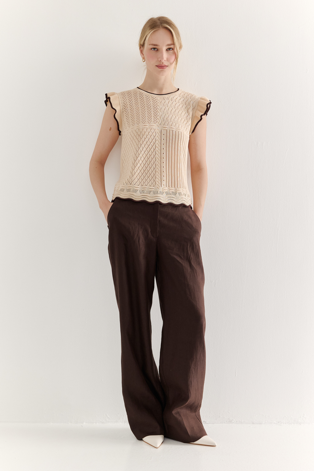 Кремовый топ в стиле пэчворк, в котором соединились четыре техники ажурной вязки, круглая горловина и деликатные рюши по пройме. Контрастная отделка коричневого цвета расположена по линиям горловины, низа и краю рюши.