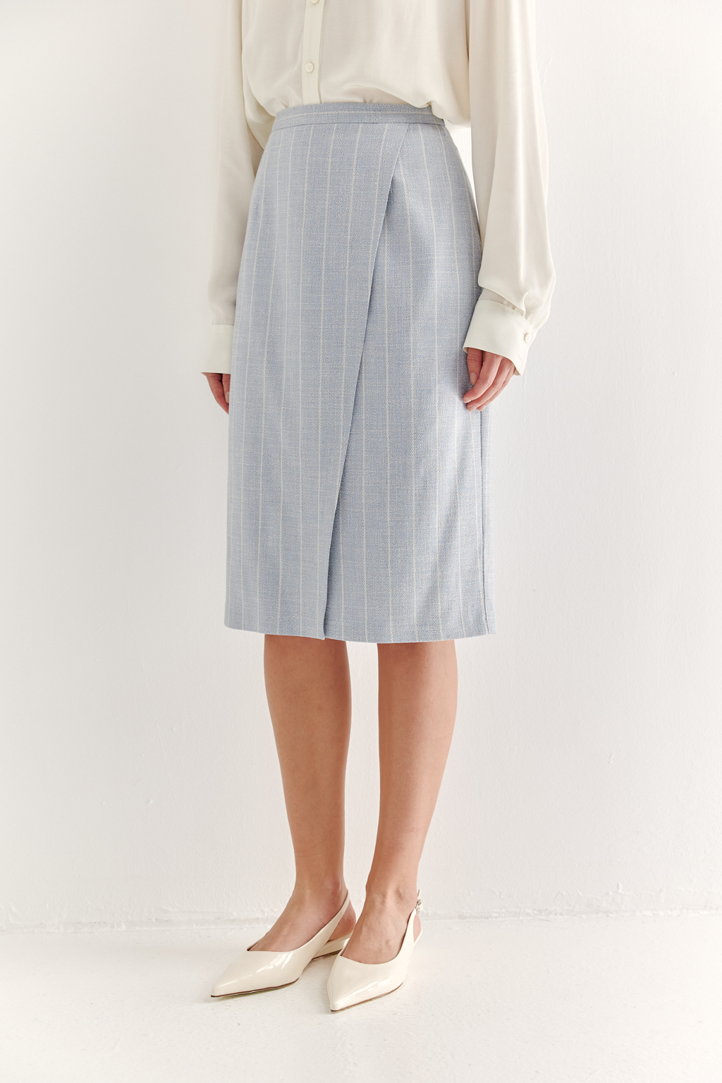 Прямая юбка с запахом выполнена из ткани, представляющей собой уникальную разработку бренда. Сочетание волокон хлопка, вискозы, полиэстера и эластана обеспечивает максимальный комфорт и практичность изделия. Нежный голубой цвет  эффектно подчёркнут светлой полоской в широком раппорте. Юбка идеально сочетается с жакетом архитектурного кроя из той же ткани, создавая завершённый формальный образ.