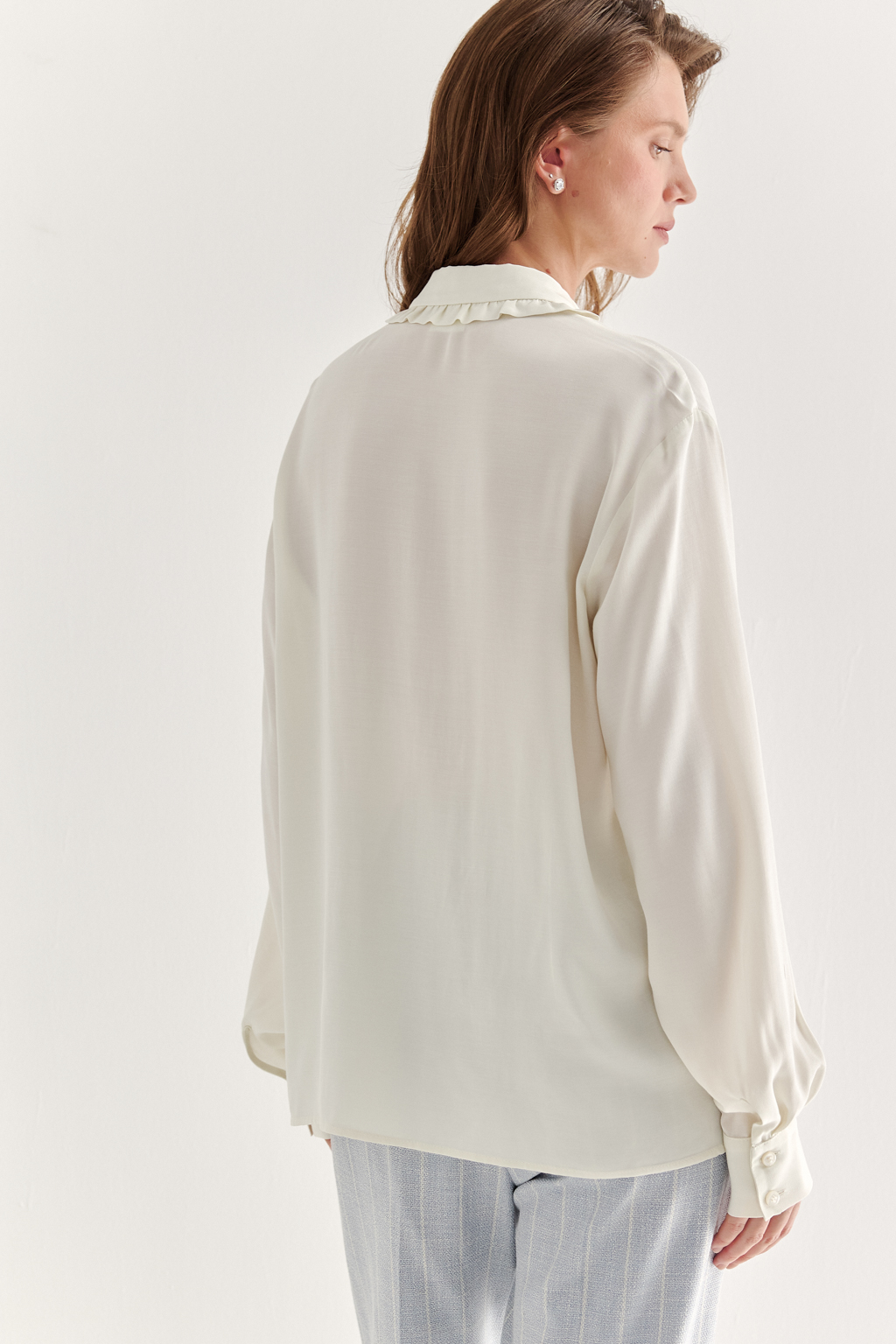 Блуза-батник в нежном оттенке сливочное масло станет изысканным дополнением к жакету, брюкам или верхней одежде, позволяя создавать актуальные многослойные образы. Изготовленная из вискозной ткани, она обладает превосходными тактильными качествами и напоминает дорогой шелковый креп. Классическая блузка свободного кроя с отложным воротником и стойкой, украшенной узким рюшем, имеет планку для застежки и втачной прямой рукав с манжетой на пуговицах.