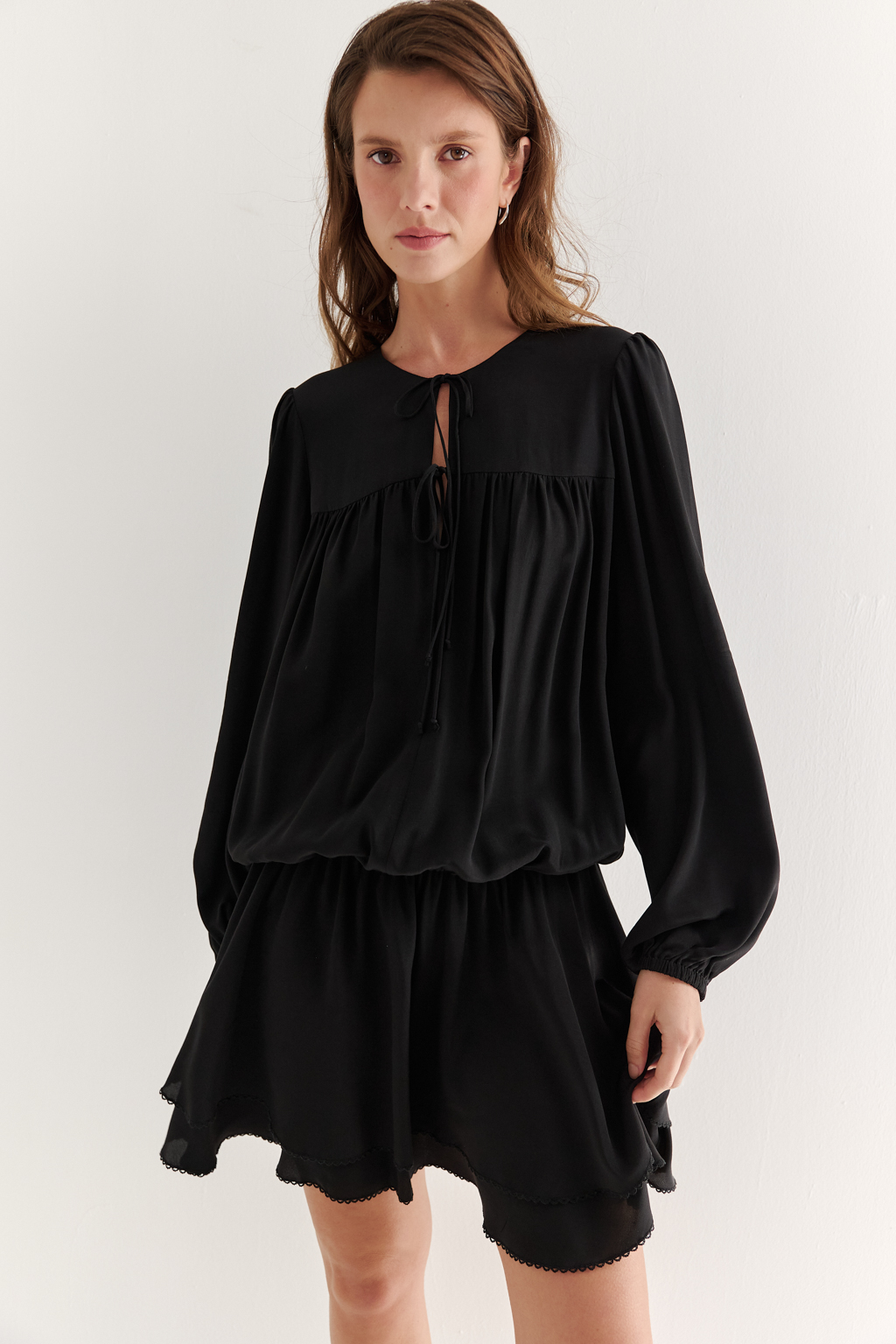 Платье, из струящейся креповой вискозы в черном цвете, представляет собой воплощение модных тенденций, умело играя с объемами с акцентом на заниженную линию талии. Это короткое, воздушное и исполненное романтики платье, сознательно избегающее вычурных деталей, является идеальным решением для создания актуального летнего городского образа. Объем платья образуется благодаря изящным сборкам, расположенным по кокеткам переда и спинки. Отрезная юбка, расширяющаяся к низу, украшена деликатной декоративной строчкой-вышивкой в виде фестонов. Линия отреза юбки оформлена эластичной тесьмой-стрейч, что дает возможность регулировать длину. Платье гармонично сочетается с рубашками или жакетами, предоставляя свободу для экспериментов со стилем. При желании можно усложнить образ образа с помощью леггинсов и также подчеркнуть талию элегантным кожаным ремнем.