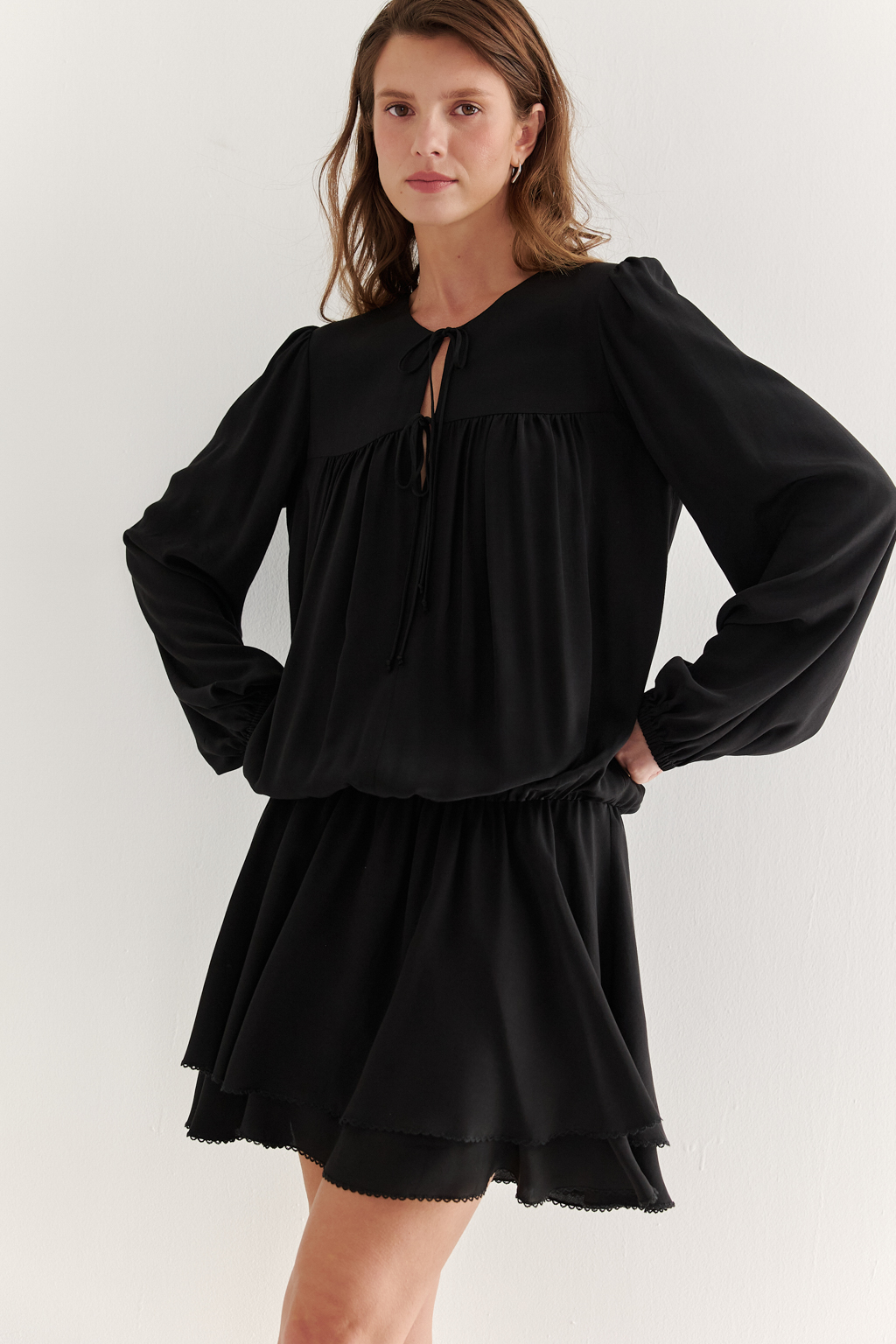 Платье, из струящейся креповой вискозы в черном цвете, представляет собой воплощение модных тенденций, умело играя с объемами с акцентом на заниженную линию талии. Это короткое, воздушное и исполненное романтики платье, сознательно избегающее вычурных деталей, является идеальным решением для создания актуального летнего городского образа. Объем платья образуется благодаря изящным сборкам, расположенным по кокеткам переда и спинки. Отрезная юбка, расширяющаяся к низу, украшена деликатной декоративной строчкой-вышивкой в виде фестонов. Линия отреза юбки оформлена эластичной тесьмой-стрейч, что дает возможность регулировать длину. Платье гармонично сочетается с рубашками или жакетами, предоставляя свободу для экспериментов со стилем. При желании можно усложнить образ образа с помощью леггинсов и также подчеркнуть талию элегантным кожаным ремнем.