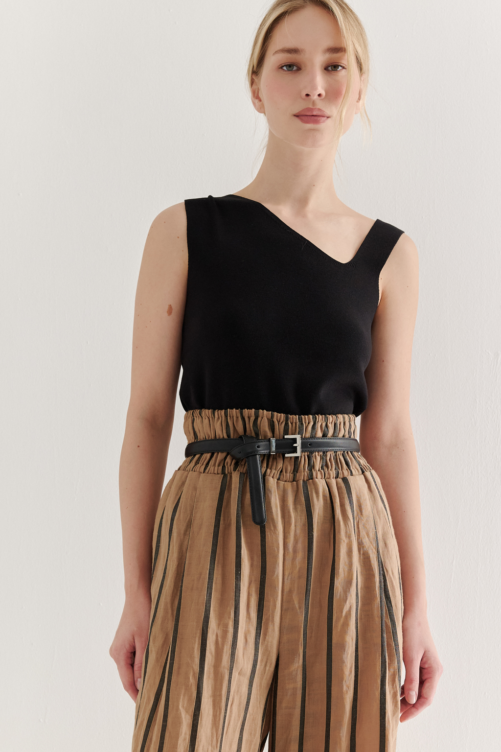 Брюки прямого кроя с широким поясом, собранным в сборку, сшиты из платьевой ткани в тонкую полоску. Состав ткани - сочетание волокон крапивы и хлопка, обработанных с эффектом растительного окрашивания (natural dye). Темная полоска на бежевом фоне идеально гармонирует с силуэтом широких брюк, подчеркивая их элегантную простоту. Пояс брюк широкий, собран на три ряда кулисок с резинками, что позволяет регулировать его высоту посадки относительно линии талии. Ткань обладает свойством создавать естественный крэш-эффект, придающий изделию легкую текстурную небрежность и объем, при этом брюки не требуют сложного ухода.