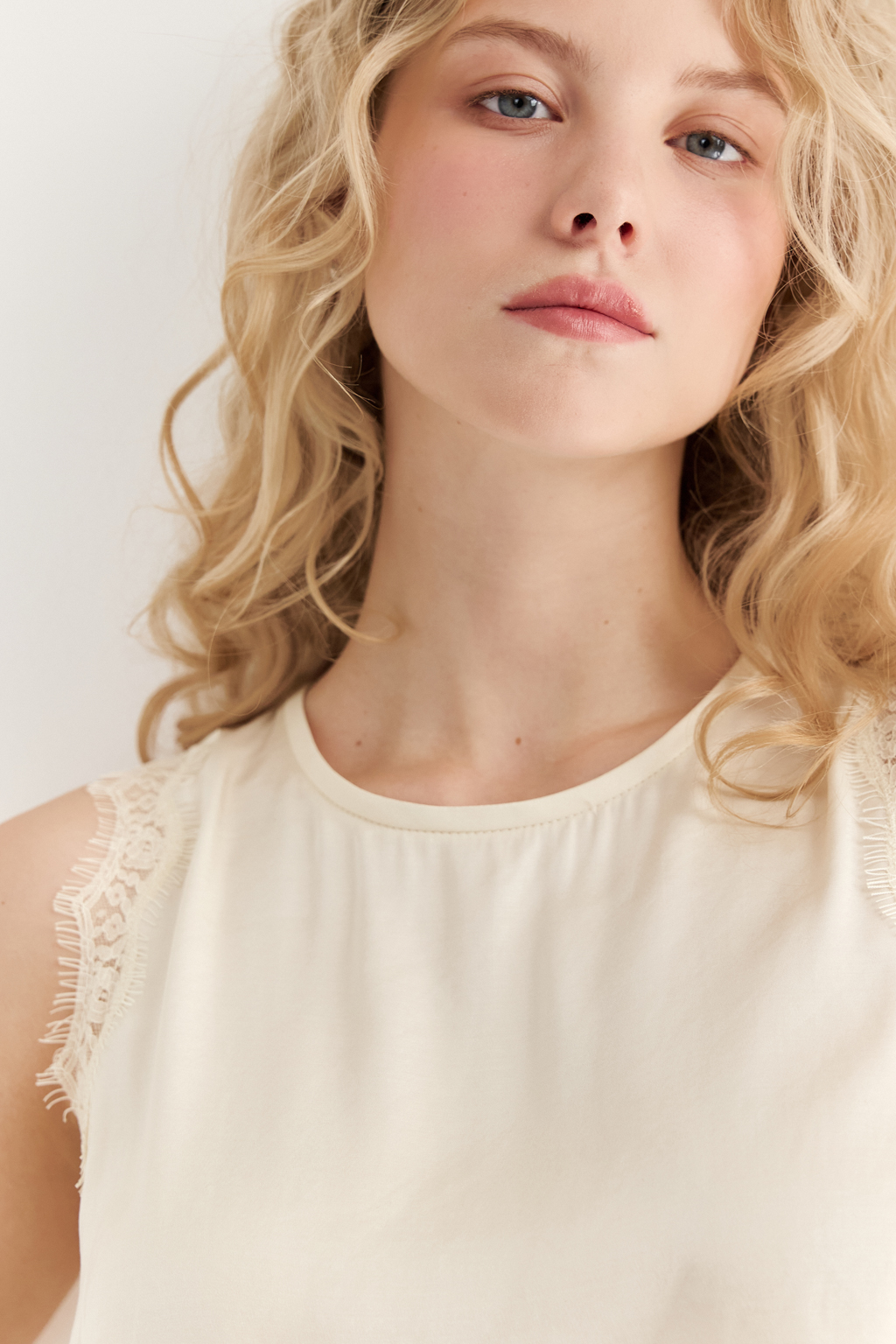 Блуза без рукавов из струящегося сатина в оттенке сливочное масло. Круглая горловина обработана кантом, на спинке воздушная петля с застежкой на пуговицу, по проймам настрочена тесьма из гипюра в тон основной ткани.