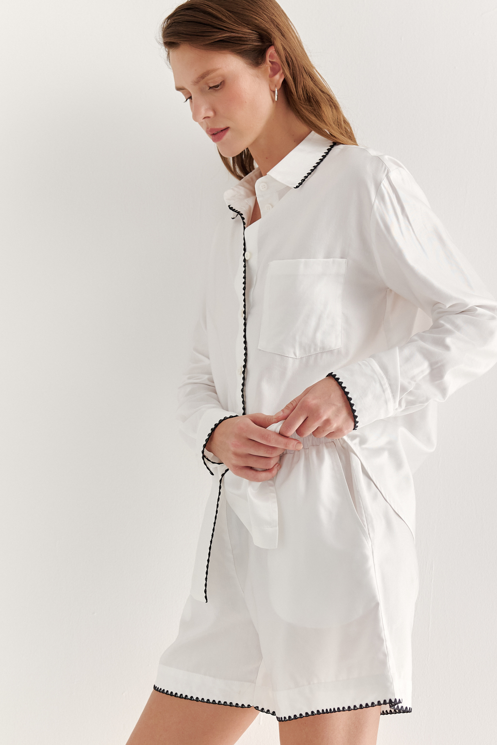 Белая блуза из нежной вискозы и дышащего хлопка, воплощает элегантность и непринужденность. Классический крой, отсылающий к мужской рубашке, характеризуется рубашечным воротником со стойкой, застежкой на пуговицы, прямыми рукавами и накладным карманом, а приподнятый у боковых швов низ придает ей легкую динамичность. Уникальность блузе придает отделка петельным декоративным швом контрастного цвета, проложенным вдоль воротника, борта и манжет. Блуза безупречно дополнит многослойный ансамбль в светлых тонах или идеально сочетается с белыми шортами, декорированными аналогичным декоративным швом.