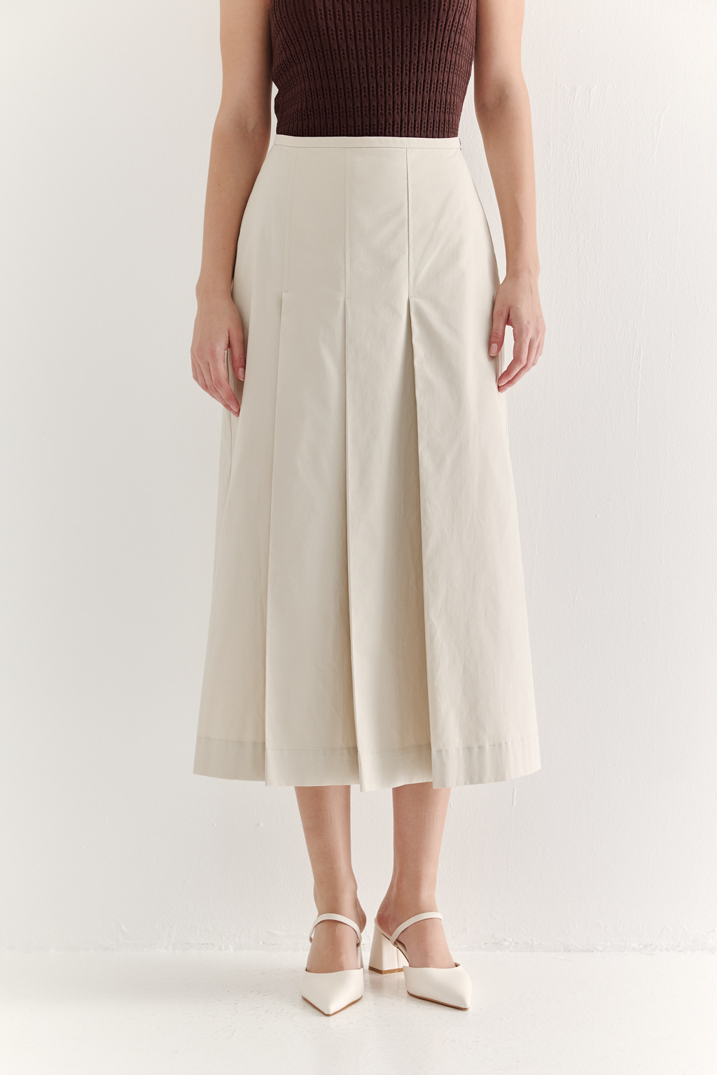Изысканная юбка-миди А-силуэта выполнена из полированного хлопка нежного светло-бежевого оттенка. Передняя часть юбки украшена тремя  складками, искусно формирующими силуэт. В крайней левой складке расположена шлица, прикрывающая высокий разрез.