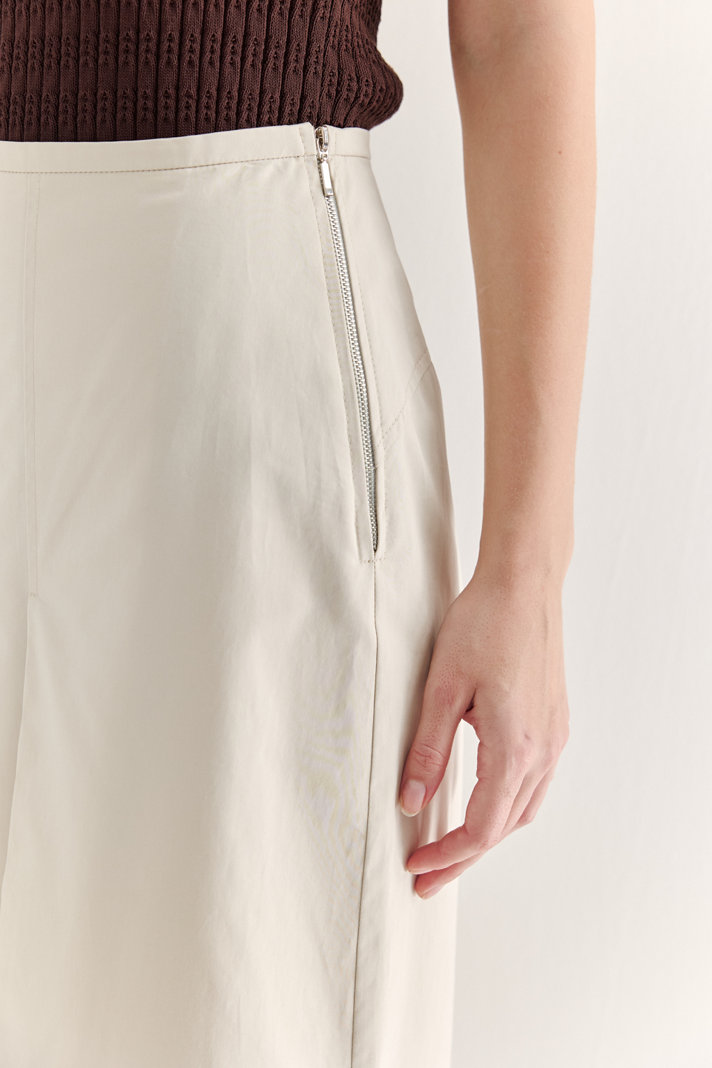 Изысканная юбка-миди А-силуэта выполнена из полированного хлопка нежного светло-бежевого оттенка. Передняя часть юбки украшена тремя  складками, искусно формирующими силуэт. В крайней левой складке расположена шлица, прикрывающая высокий разрез.