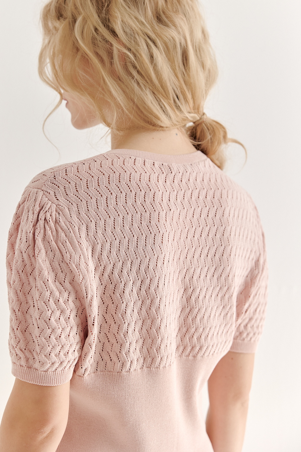 Джемпер нежно-розового цвета, связанный из тонкой тактильной пряжи, в составе которой вискоза и нейлон. В дизайне джемпера применен принцип контраста фактур, в данном случае – полупрозрачного ажура и ластика. Круглая горловина и планка с застежкой на 9 пуговиц-жемчужин, рукав длиной одна вторая.