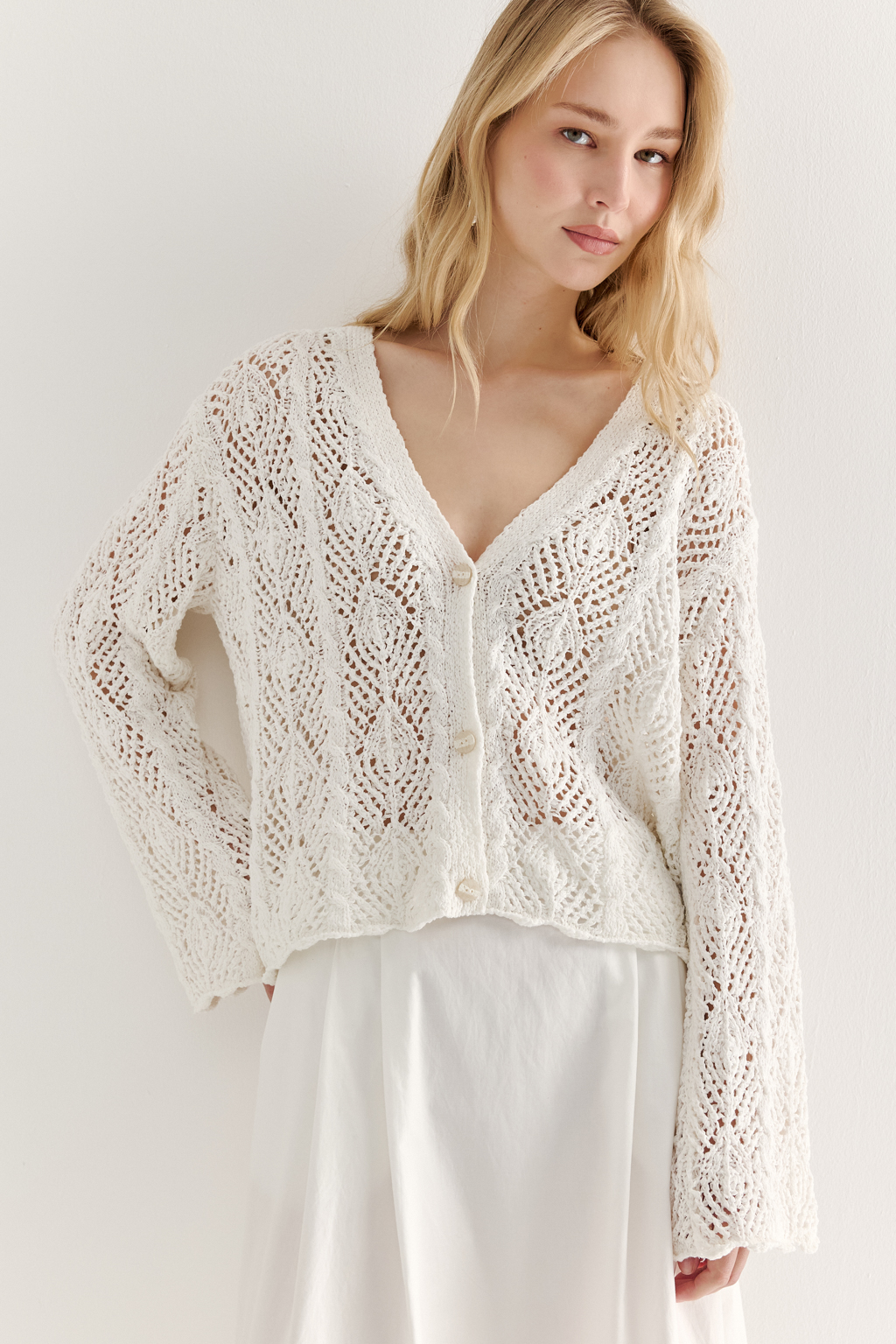 Белый вязаный кардиган оверсайз силуэта из хлопковой пряжи. Кардиган связан в сложной технике комбинирования сетки и ажурных ромбов. Рукав прямой удлиненный, V-образный вырез, борт отделан планкой, застежка по борту на 3 пуговицы, низ кардигана и рукав – сложные фестоны.