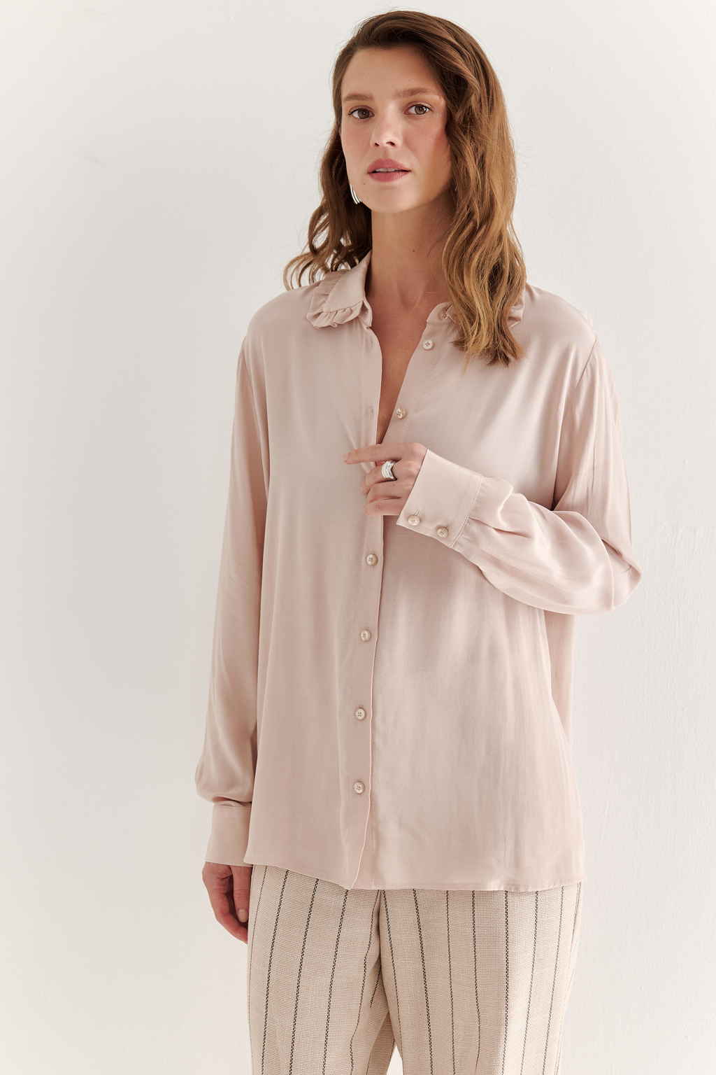 Блуза-батник в нежном пудрово-розовом оттенке станет изысканным дополнением к жакету, брюкам или верхней одежде, позволяя создавать актуальные многослойные образы. Изготовленная из вискозной ткани, она обладает превосходными тактильными качествами и напоминает дорогой шелковый креп. Классическая блузка свободного кроя с отложным воротником и стойкой, украшенной узким рюшем, имеет кокетку спинки, планку для застежки и втачной прямой рукав с манжетой на пуговицах.