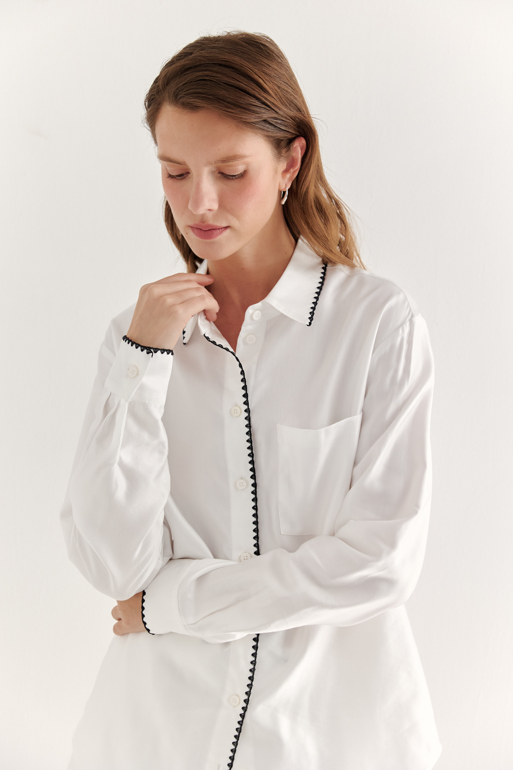 Белая блуза из нежной вискозы и дышащего хлопка, воплощает элегантность и непринужденность. Классический крой, отсылающий к мужской рубашке, характеризуется рубашечным воротником со стойкой, застежкой на пуговицы, прямыми рукавами и накладным карманом, а приподнятый у боковых швов низ придает ей легкую динамичность. Уникальность блузе придает отделка петельным декоративным швом контрастного цвета, проложенным вдоль воротника, борта и манжет. Блуза безупречно дополнит многослойный ансамбль в светлых тонах или идеально сочетается с белыми шортами, декорированными аналогичным декоративным швом.