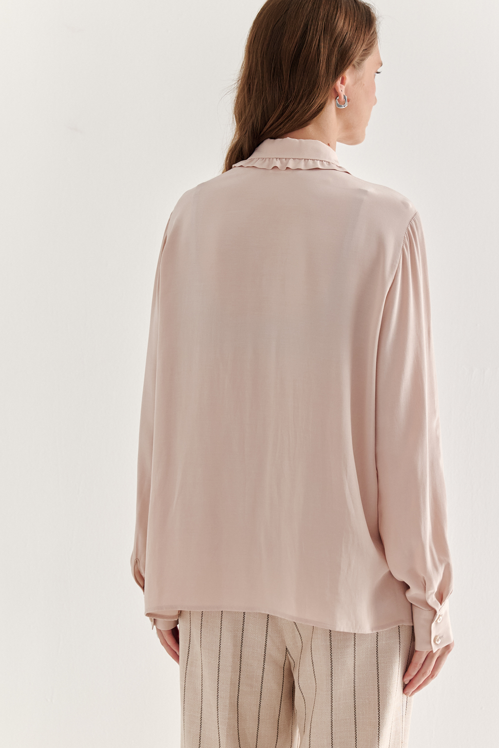 Блуза-батник в нежном пудрово-розовом оттенке станет изысканным дополнением к жакету, брюкам или верхней одежде, позволяя создавать актуальные многослойные образы. Изготовленная из вискозной ткани, она обладает превосходными тактильными качествами и напоминает дорогой шелковый креп. Классическая блузка свободного кроя с отложным воротником и стойкой, украшенной узким рюшем, имеет кокетку спинки, планку для застежки и втачной прямой рукав с манжетой на пуговицах.