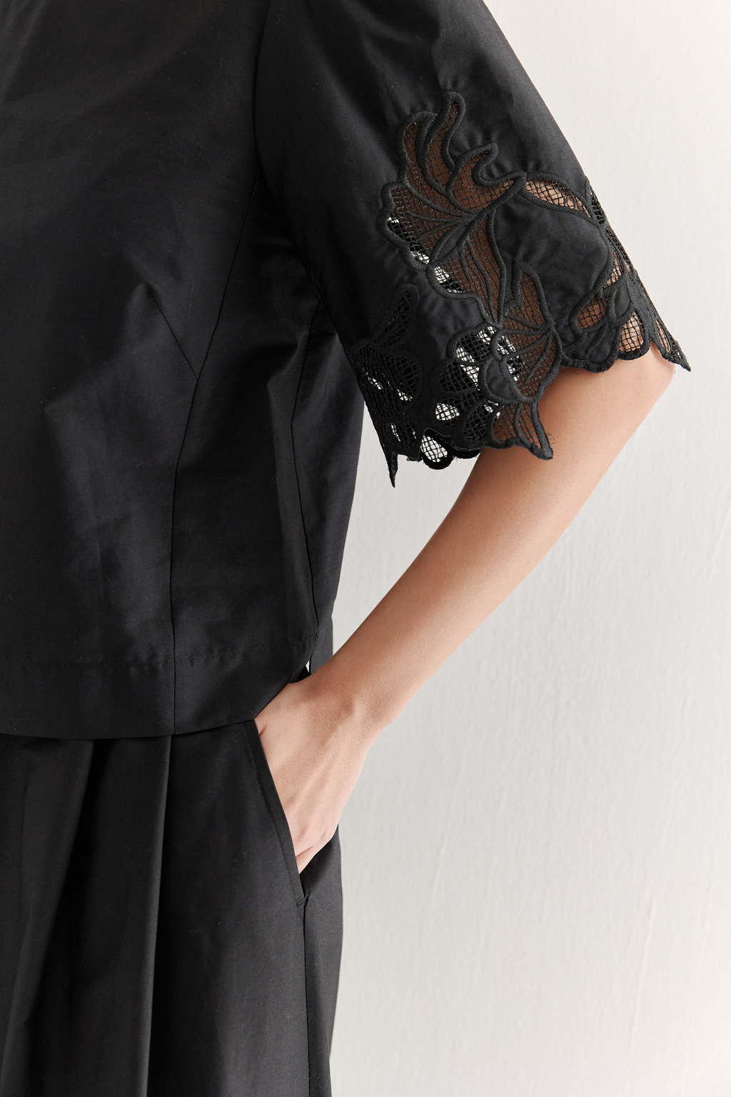 Блуза-кроп выполненная из хлопкового полотна, декорирована нежным шитьем-решелье черного цвета. Укороченный силуэт блузы дополнен широкой подгибкой по низу и элегантными разрезами в боковых швах. Горловина оформлена в виде лодочки, визуально удлиняя шею. Широкие рукава имеют сборку по окату и украшены шитьем по низу, что придает образу легкости и женственности. Молния, расположенная в среднем шве спинки, обеспечивает комфорт и удобство при носке. Идеально подойдет к юбке с шитьем-решелье белого цвета.