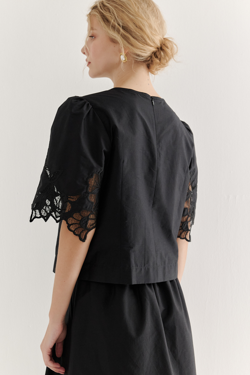 Блуза-кроп выполненная из хлопкового полотна, декорирована нежным шитьем-решелье черного цвета. Укороченный силуэт блузы дополнен широкой подгибкой по низу и элегантными разрезами в боковых швах. Горловина оформлена в виде лодочки, визуально удлиняя шею. Широкие рукава имеют сборку по окату и украшены шитьем по низу, что придает образу легкости и женственности. Молния, расположенная в среднем шве спинки, обеспечивает комфорт и удобство при носке. Идеально подойдет к юбке с шитьем-решелье белого цвета.