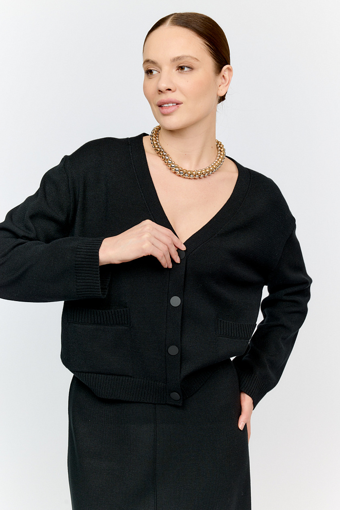 Трикотажный кардиган в базовом черном цвете с V-образным вырезом горловины, на пуговицах, со втачным рукавом,  двумя накладными карманами на полочке. Можно использовать в качестве альтернативы жакетам и носить вторым слоем в расстегнутом виде на более тонкие верха.